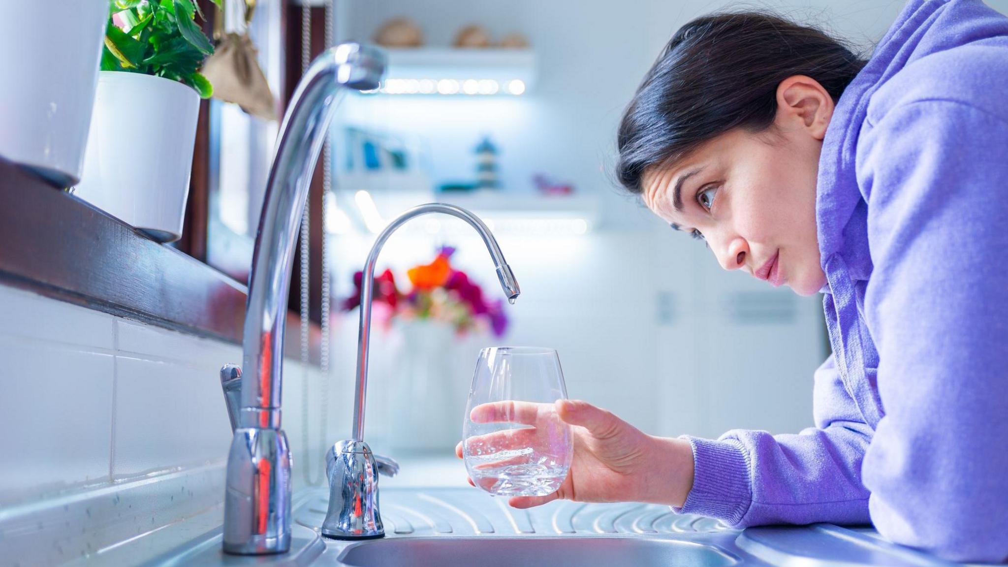 A young holds an empty glass under a tap in a kitchen.  She has dark hair tied back and is wearing a bright purple hooded top 