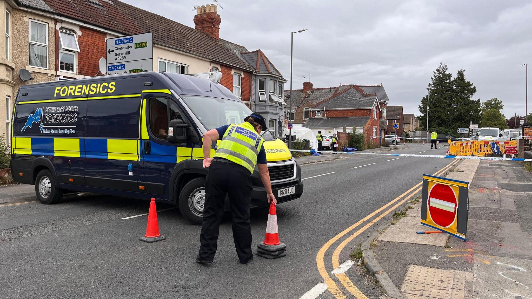 A police officer, seen only from behind, moves a traffic cone in front of a closed road. There is a large police forensics van parked across the road, as well as signs and blue police tape. A white forensics tent can be seen in the distance in front of a house.