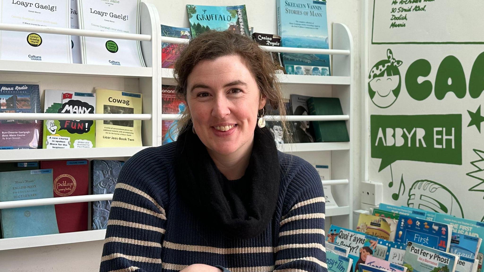 Ruth Keggin-Gell has dark brown hair tied back in a ponytail and wears a blue and cream stripped jumper and looped scarf. She is sitting in front of a bookcase containing books written in the Manx language.