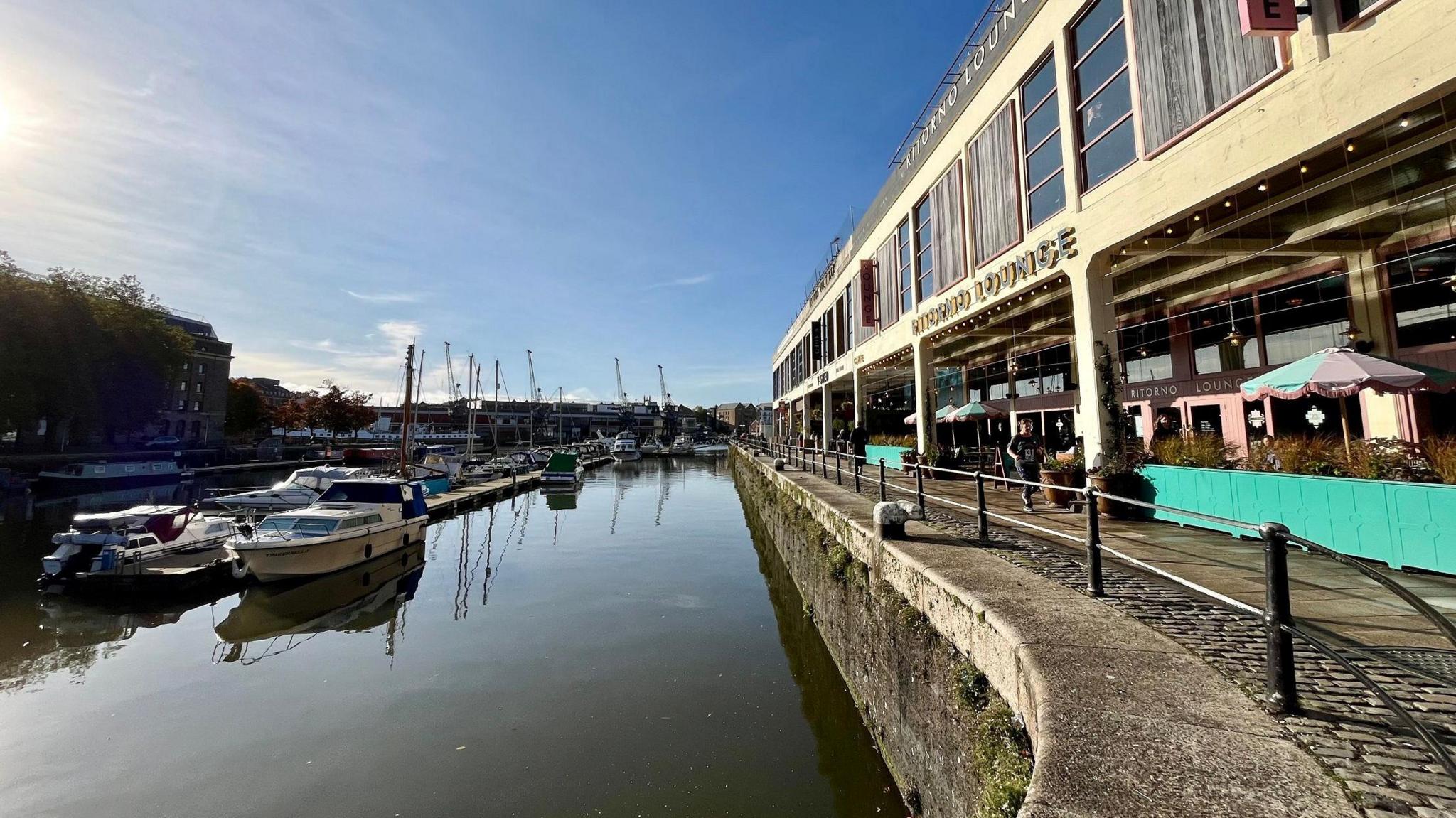 Bristol Harbourside can be seen on a sunny day, with boats moored up and bars on the side. The water is calm and the MShed Museum can be seen in the background