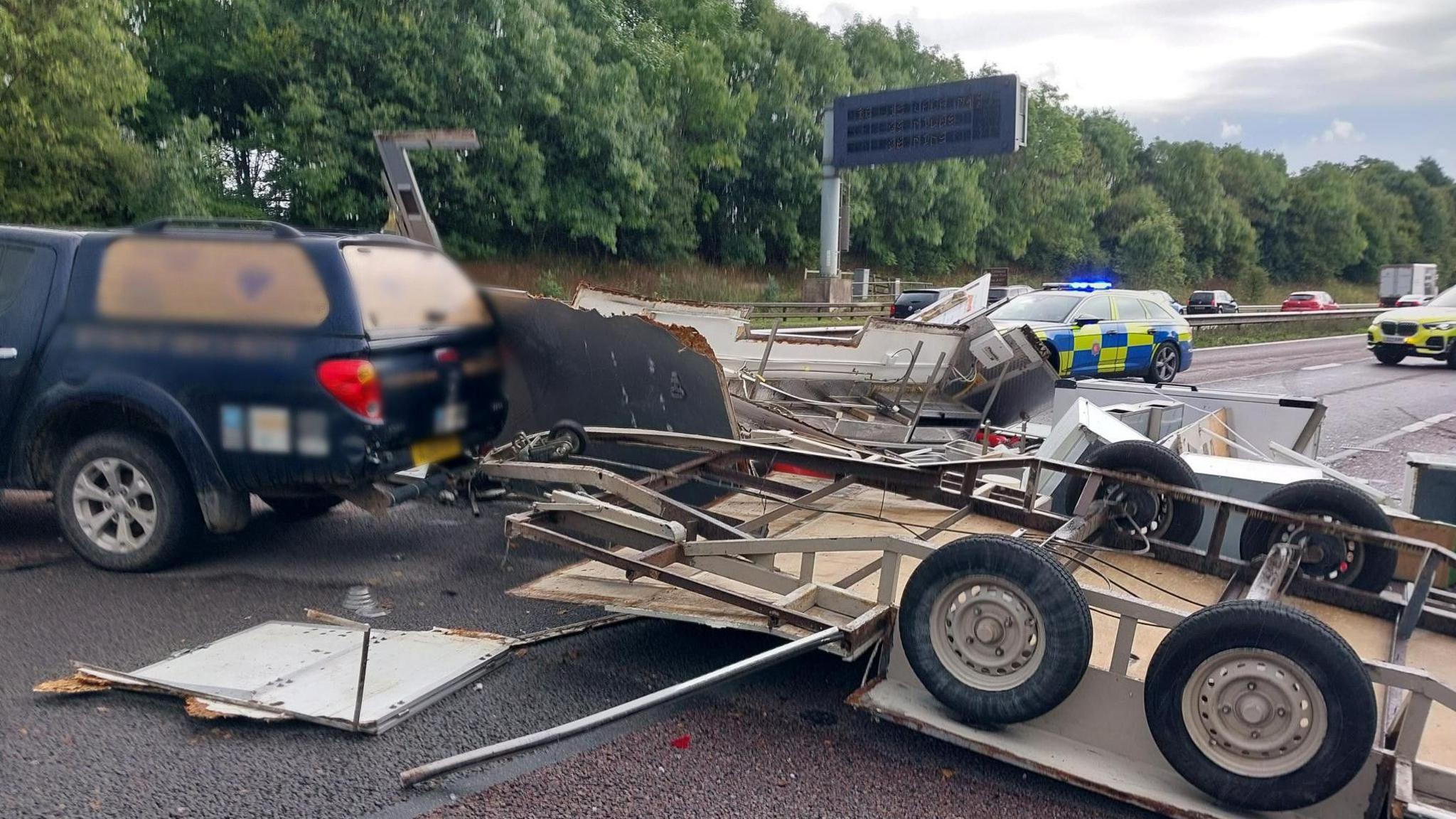 A broken up trailer can be seen having scattered white goods and what appear to be building materials. The trailer is attached to a dark-coloured 4x4 vehicle and in the background two police cars are visible with their blue lights on. 