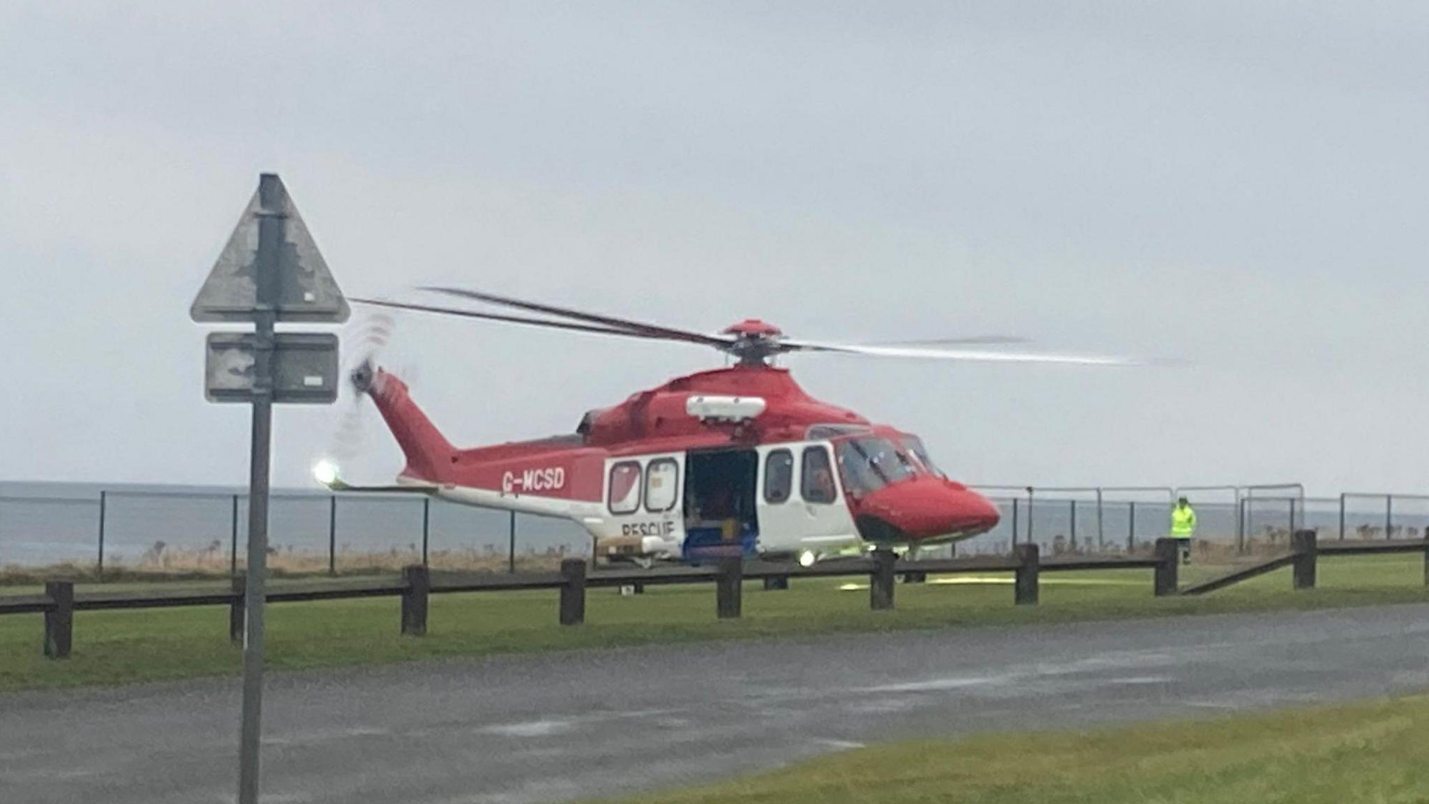 A red and white helicopter on grass in front of a fence and the North Sea. A person in a green hi-vis jacket is stood the the side of the helicopter. 