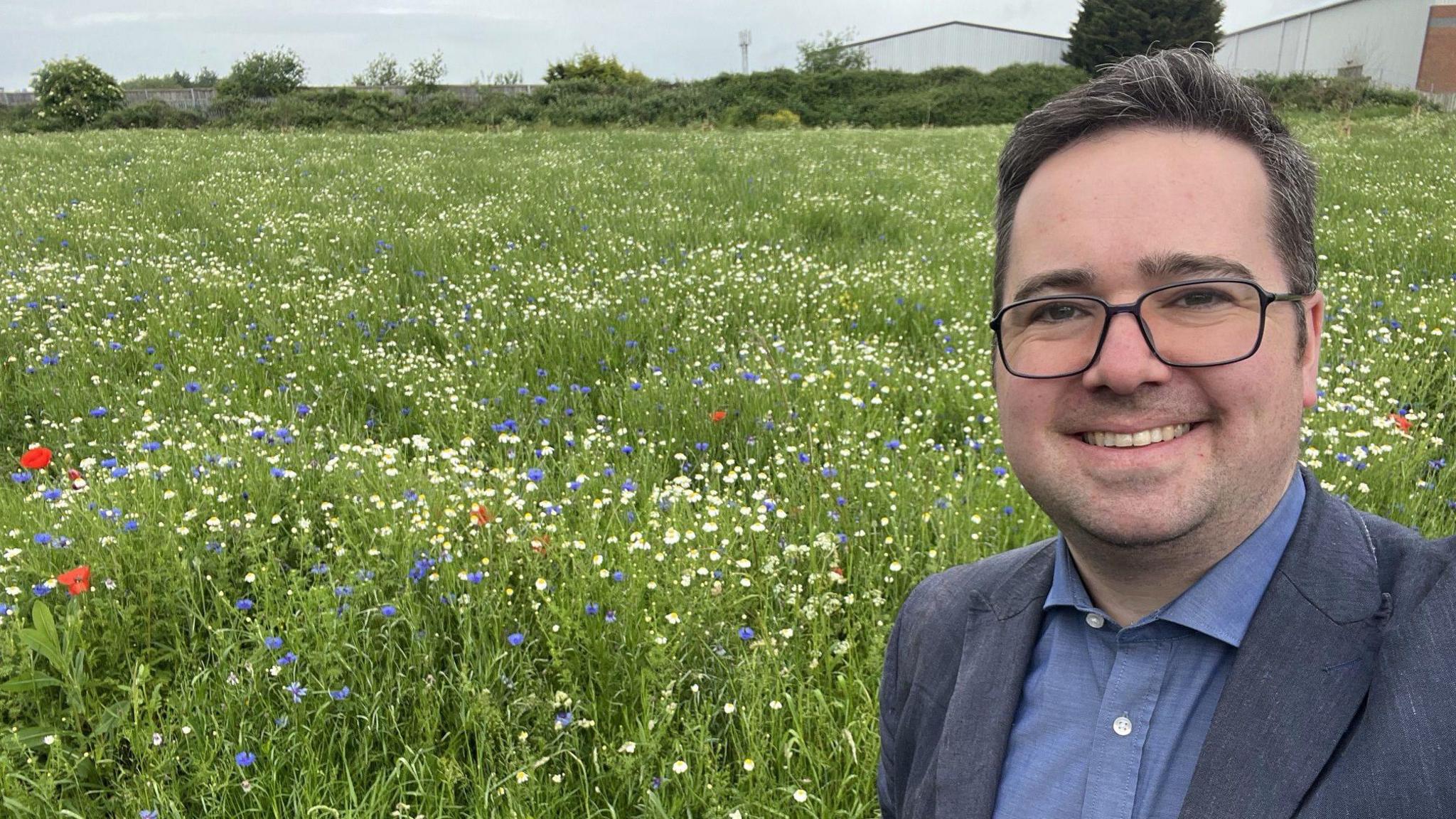 Councillor Sebastian Field - a middle-aged man with short dark greying hair wearing black rimmed glasses and a grey suit with a blue shirt - stands in the middle of a field full of wildflowers, smiling.