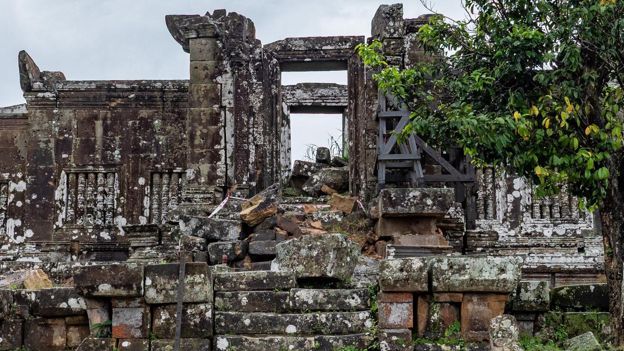Stone stairways leading up to an ancient temple appear damaged, with some parts of it having collapsed. A tree stands next to the entrance of the temple.  