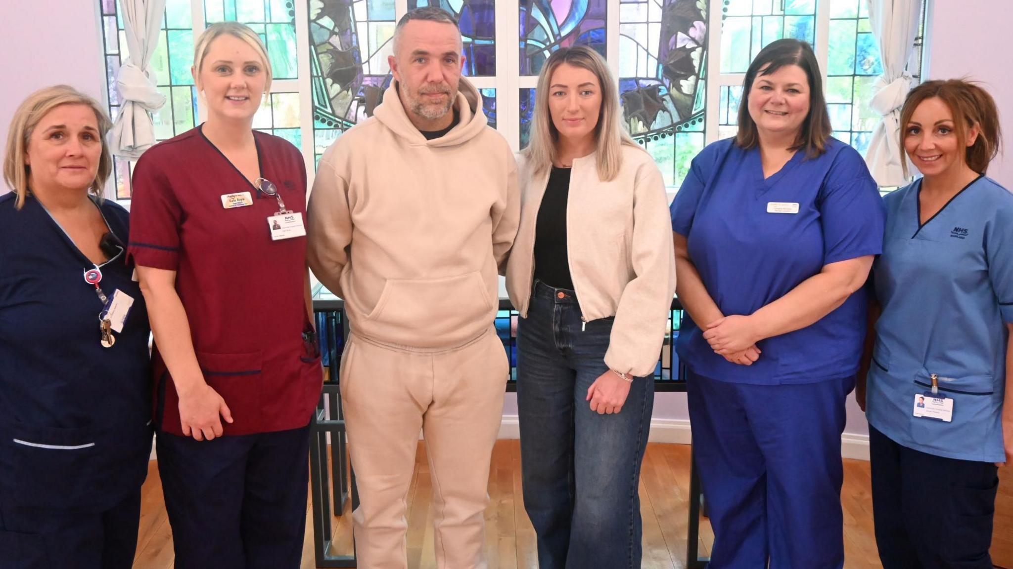 A man with short cropped hair and navy grey stubble stands next to a woman with blonde hair. She is wearing a white jacket, black top and blue jeans. Two female members - both with brown hair- of the nursing staff are standing to the woman's left wearing blue hospital overalls. Two blonde female members of the nursing staff are standing to the right of the man. One is wearing a maroon NHS top with black trousers while the other is wearing a navy blue uniform. The couple have serious facial expressions but the staff members are smiling. A stained glass window can be seen in the background.