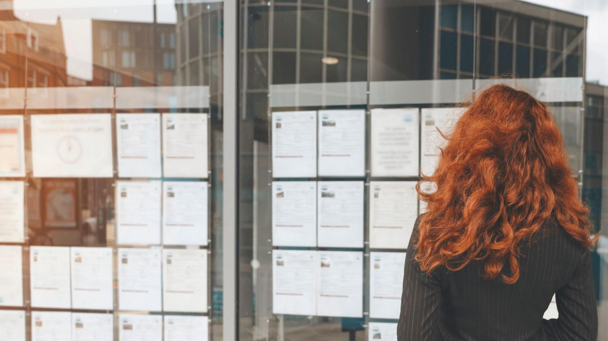 A woman with long red hair stands in front of a bulletin board with job listings, reviewing the possibilities.