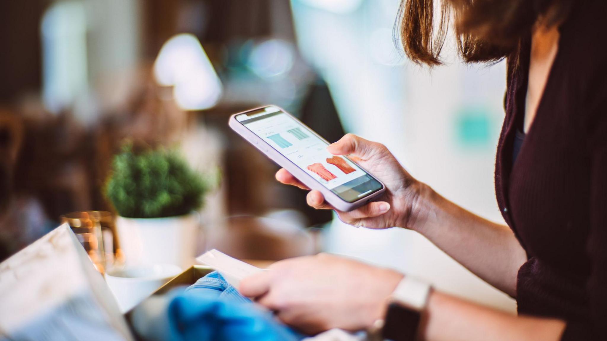 Woman holds phone checking clothes online as she stands in a shop with the item.