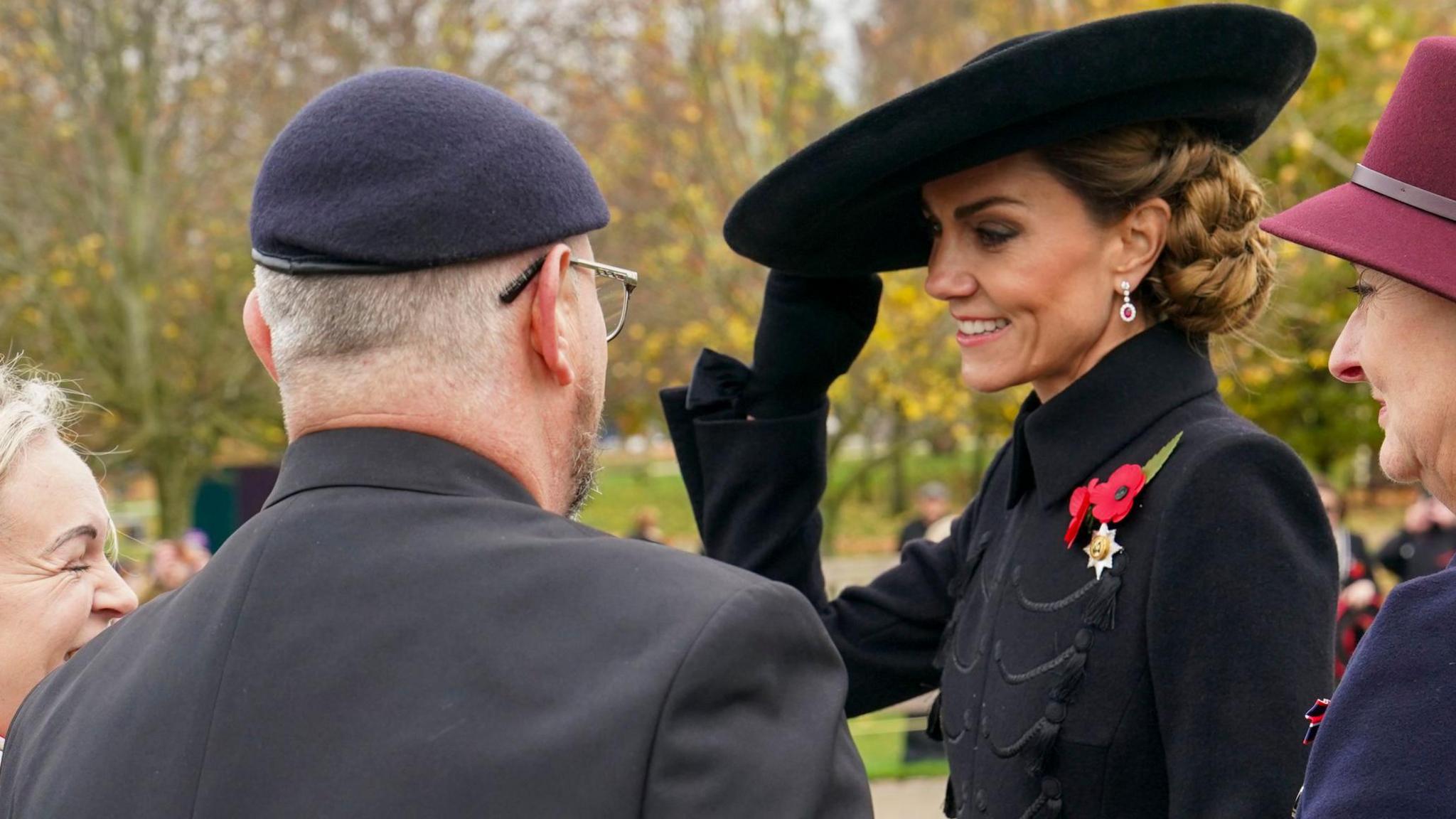 The Princess of Wales is standing in front of one man and two women. She is holding a large black hat on her head and wearing black gloves, a long black coat with two red poppies on her left chest and her brown hair is tied up. The man has his back to the camera and is wearing a black suit, navy beret and glasses. The woman to the right is wearing a maroon hat and a navy coat. The lady on the left has long blonde hair, a black coat and a scarf decorated with poppies. There are lots of blurred out trees behind them.