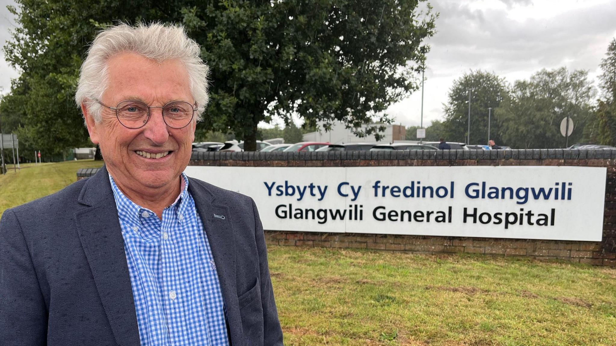 Gareth John smiles at the camera standing in front of a low brick wall with a white sign on in that reads 'Glangwili General Hospital' in both Welsh and English. He is wearing a grey blazer jacket and a blue and white chequered shirt, and has glasses and short white hair.