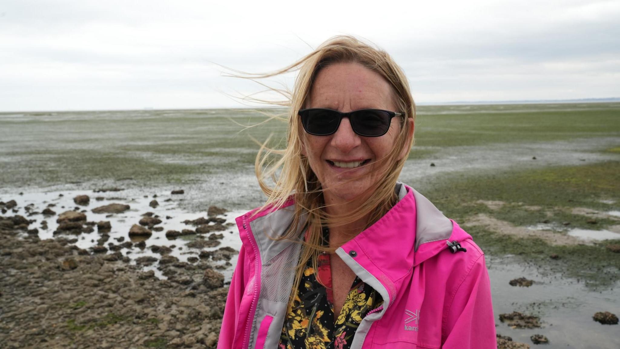 A woman, Julie-Ann Mason, wears a bright pink coat and a floral top. She is wearing sunglasses and has long blonde hair. She is smiling at the camera. A coastal landscape with green and brown hues is behind her, along with a cloudy sky. 