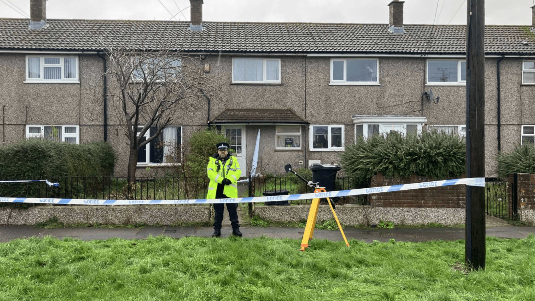 A police officer outside the address in Moredon. It is a terraced property that faces a grassy area. Police tape can be seen across the street.