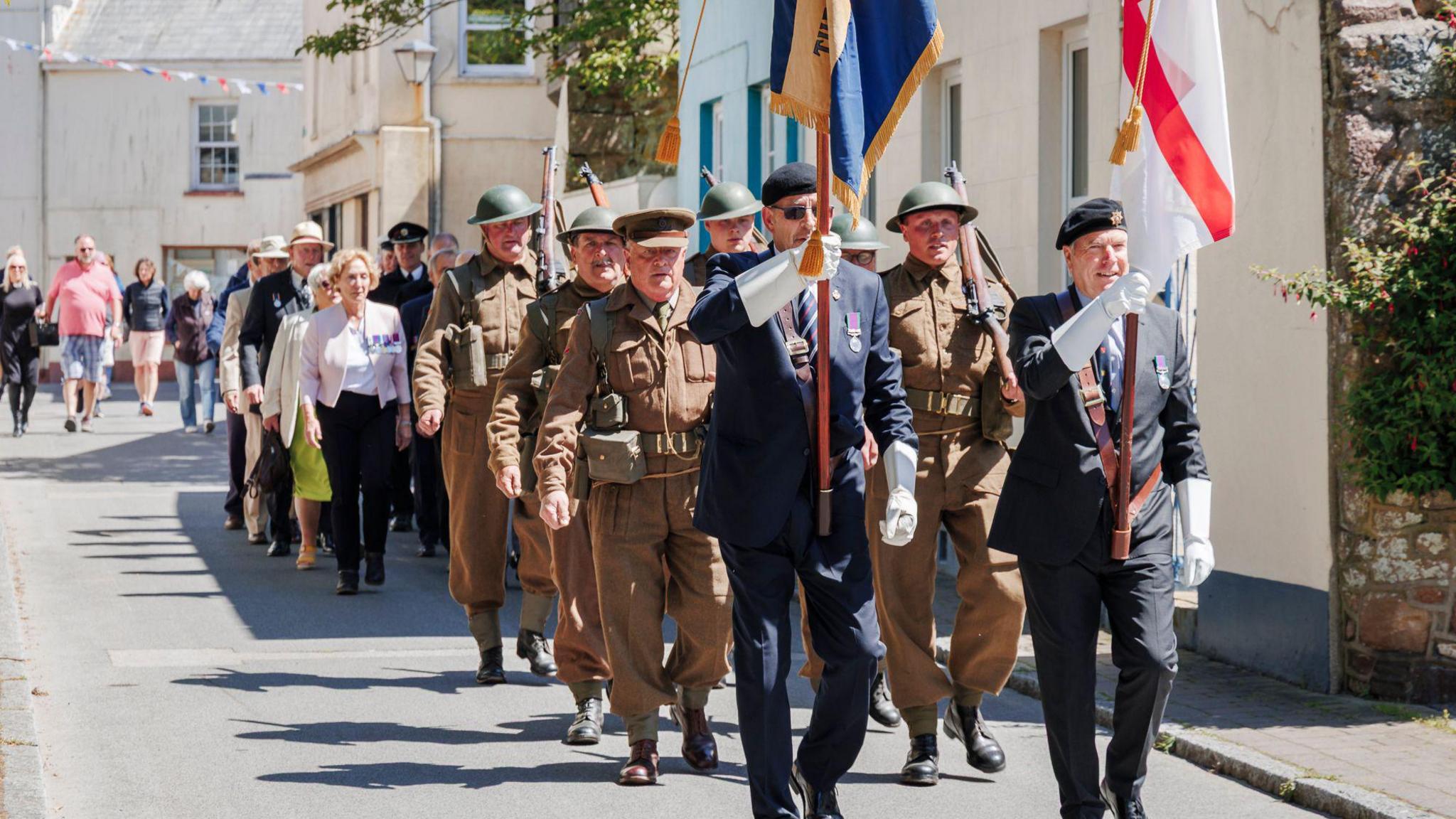 Two men in suits with war military medals over their left breast carry a large red and white flag, as well as a blue and gold flag. Soldiers in historic military uniform follow behind them. Behind the soldiers are members of the public.