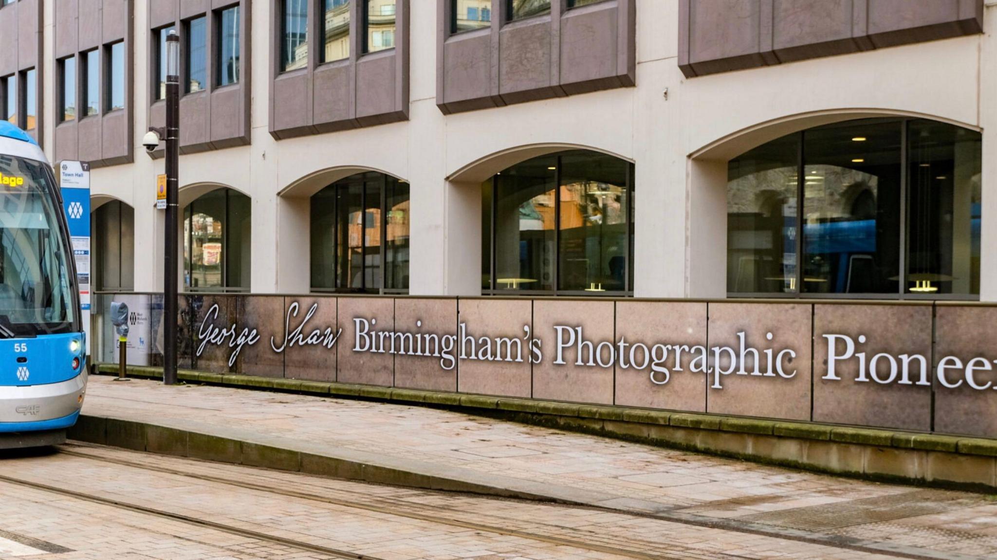 A West Midlands Metro tram is pictures to the left of a long board reading "George Shaw, Birmingham's Photographic Pioneer"