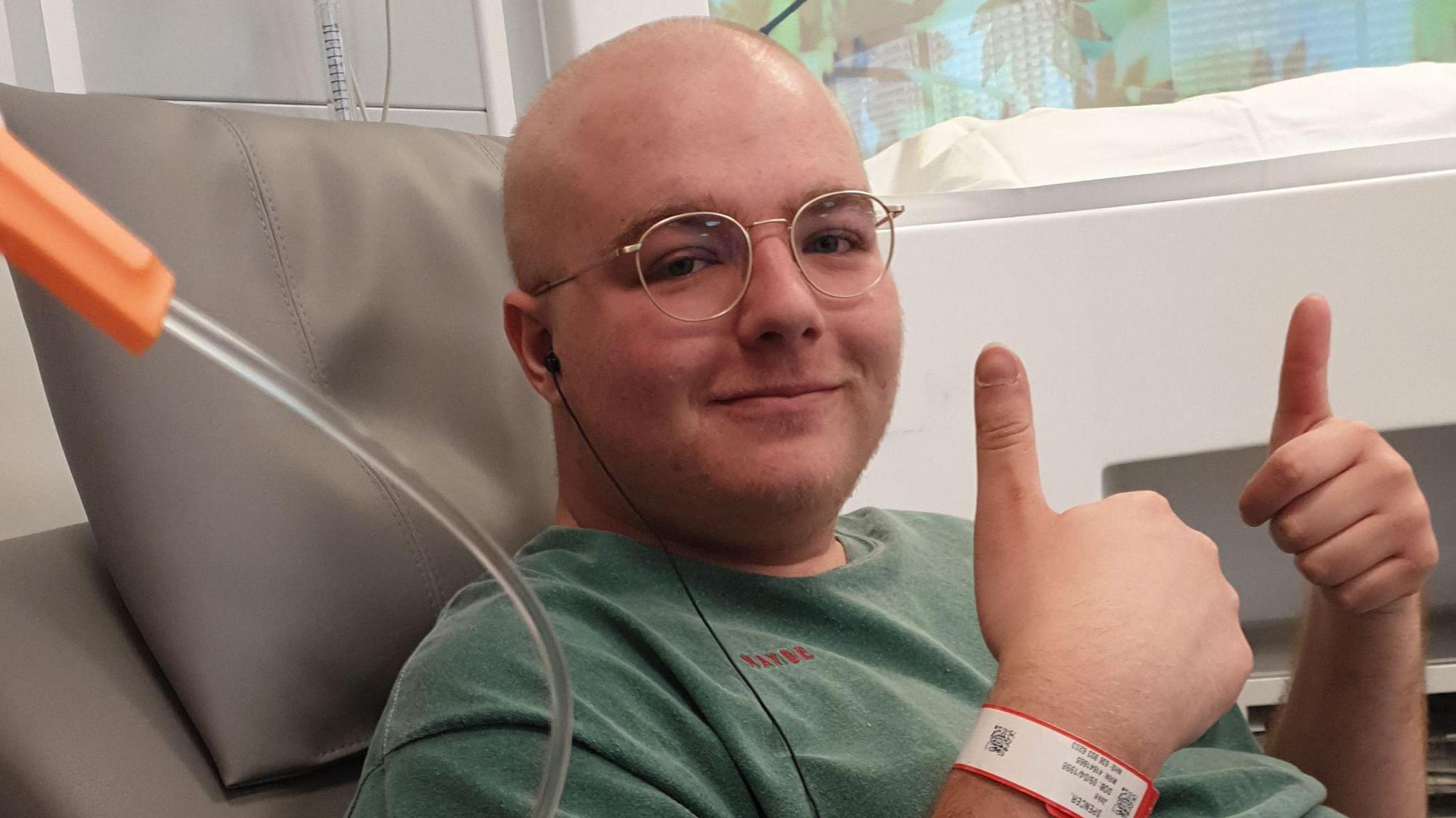 Jake Spencer sits in a hospital chair looking at the camera with two thumbs up