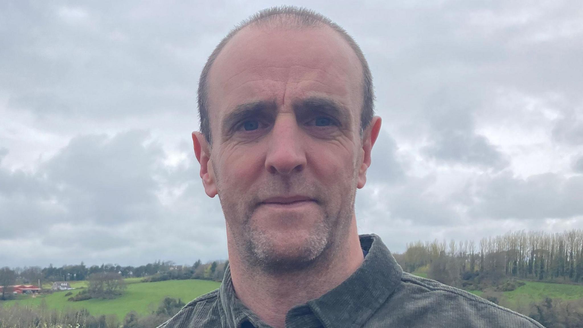 Mark H Durkan - a man wearing a green corduroy shirt looks at the camera while standing in front of farmland and a body of water.