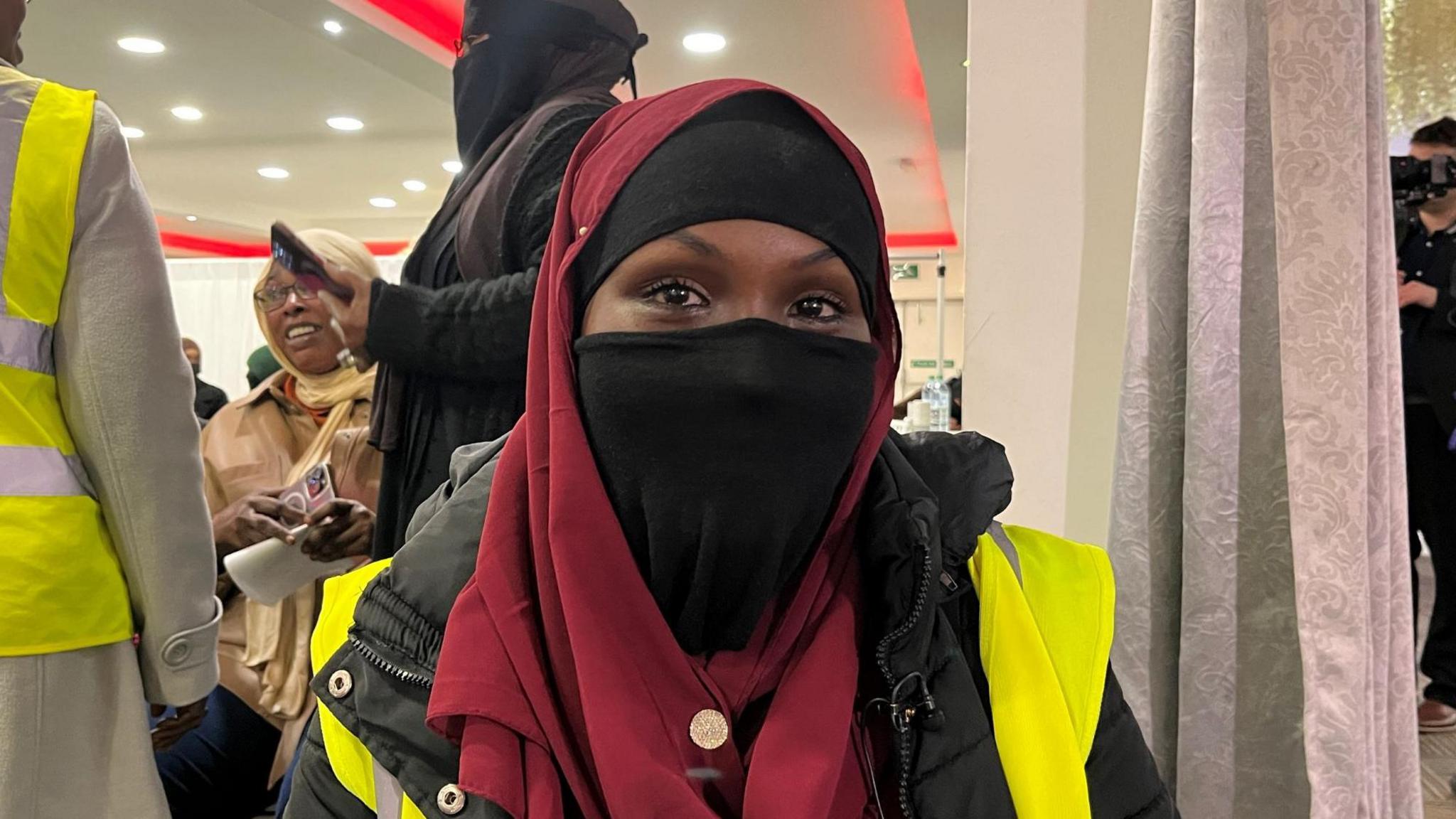 HelloFresh worker, Tasmin wearing a maroon headdress, black coat and a fluorescent jacket. Her face is covered by a black scarf below the eyes.