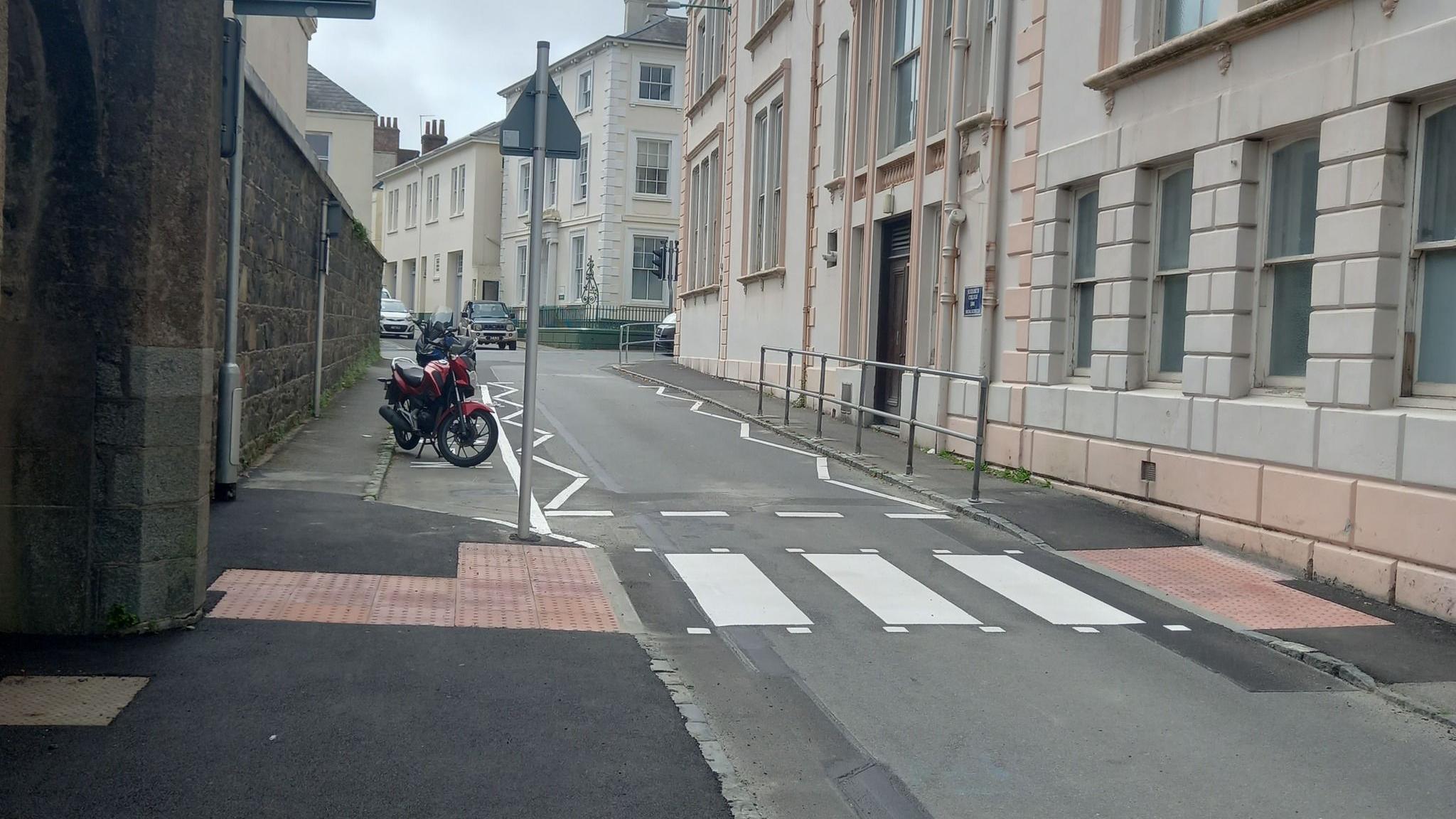 A zebra crossing on a single-lane road in Guernsey surrounded by buildings, with two cars in the distance and a parked motorbike on the left