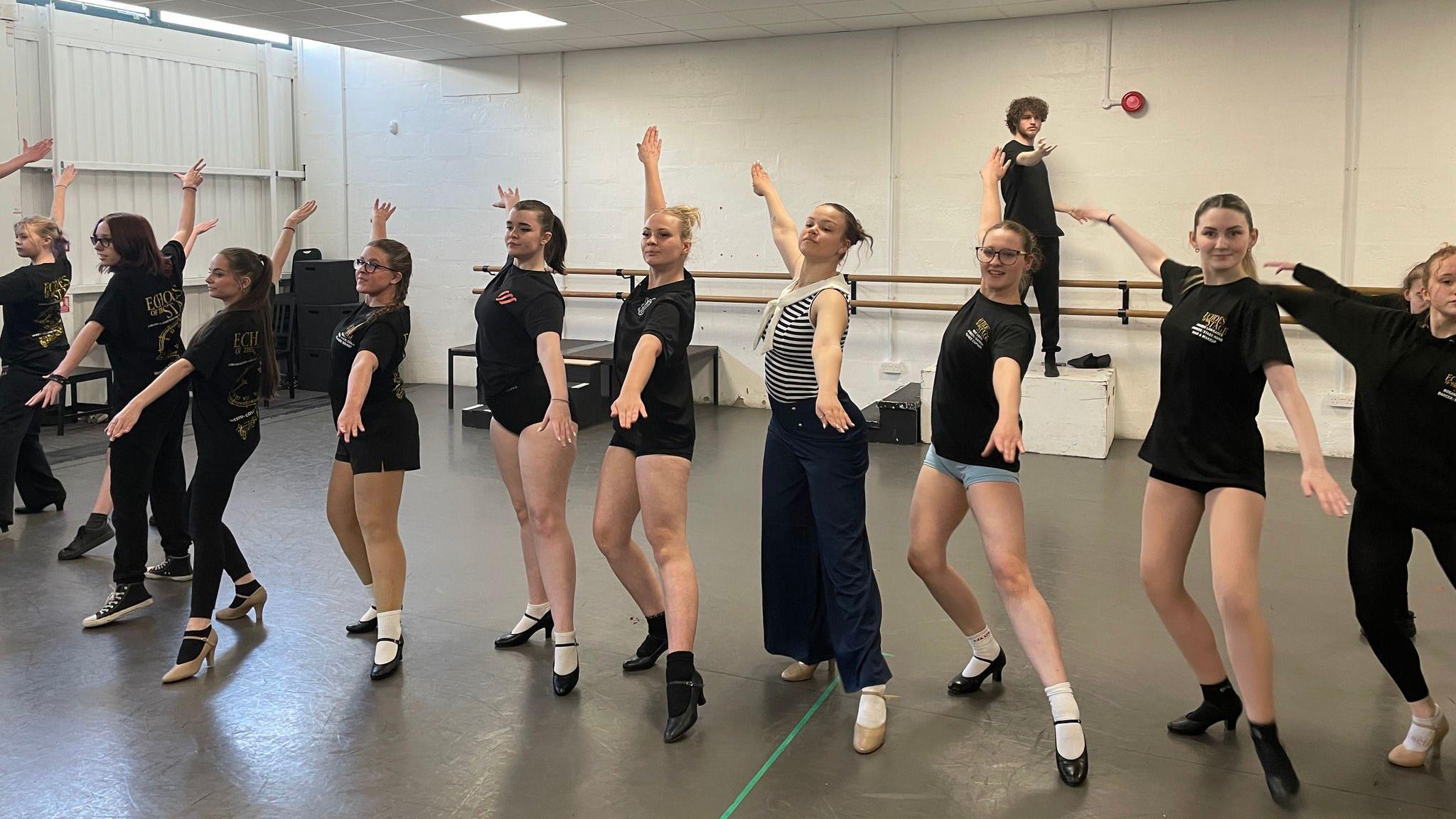 A group of young people are standing in a row with their legs and arms stretched out to practice a dance. They are in a gym.