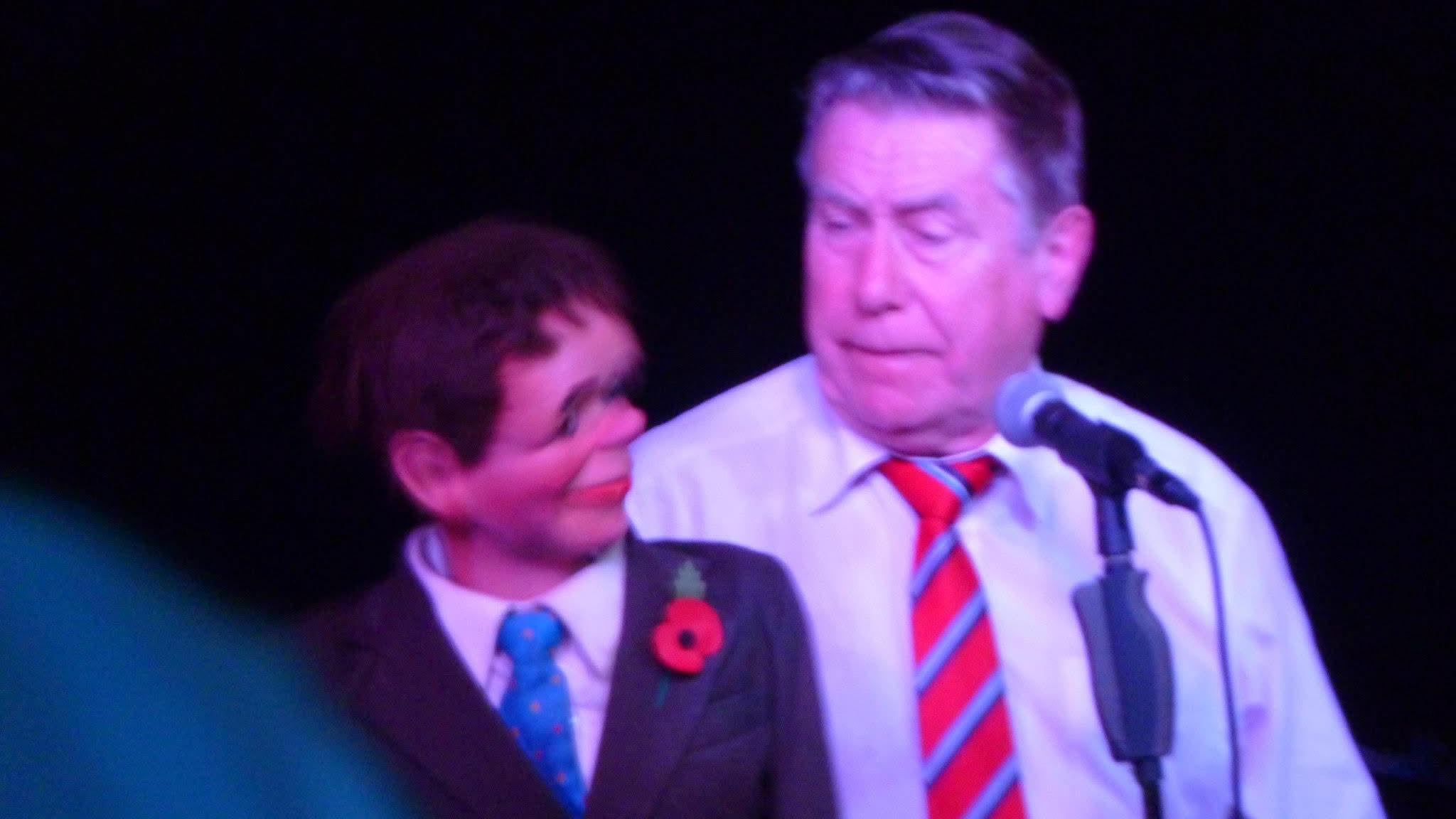 Mr Dennett is wearing a light coloured shirt with a red and blue striped tie. He is holding his ventriloquist's doll, Chic, with a microphone on a stand in front of him. Mr Dennett is looking at Chic with a concerned expression on his face. Chic is wearing a dark coloured suit with a poppy pinned to the lapel, along with a white shirt and a turquoise-coloured tie. 