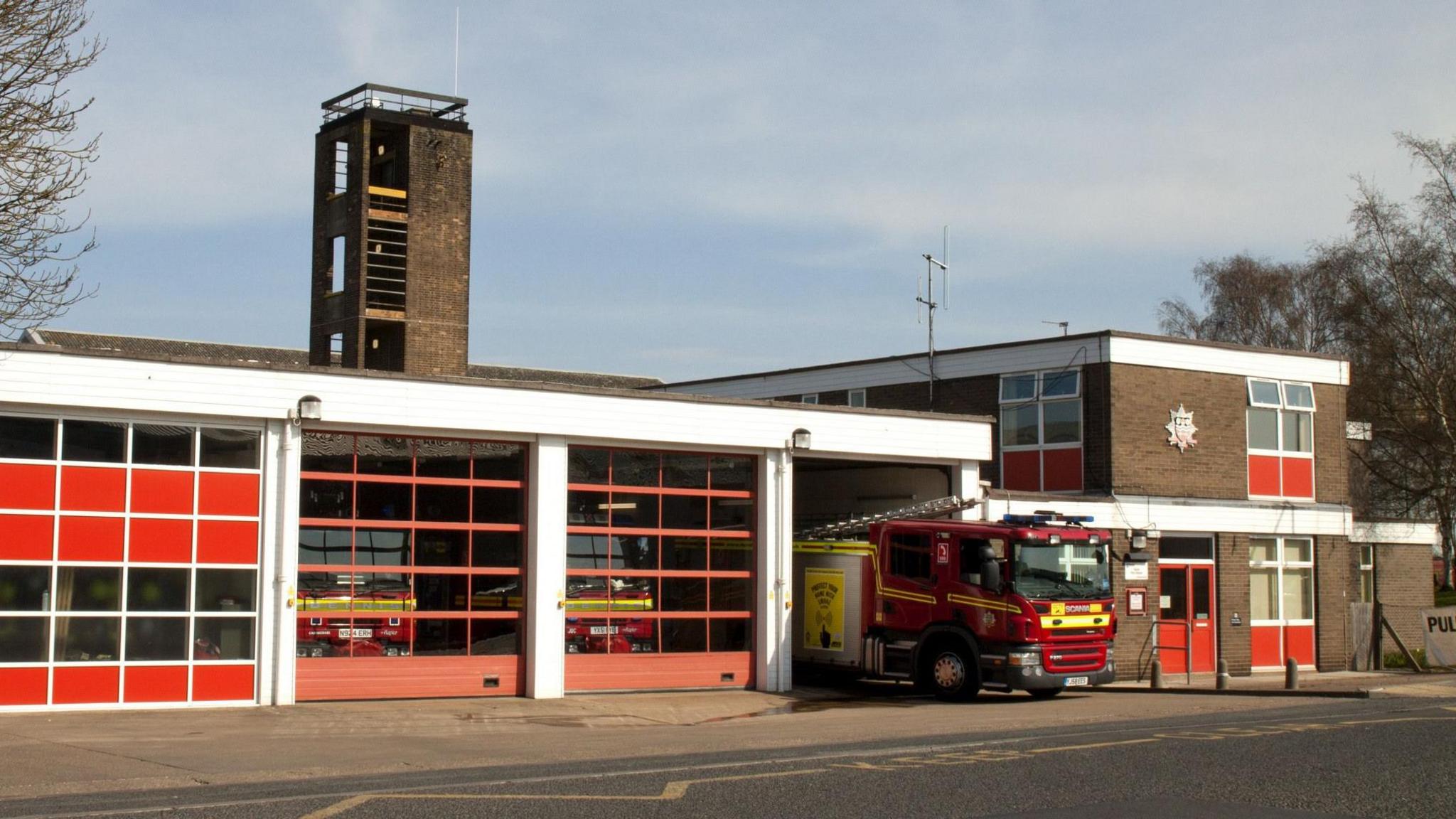 The exterior of a fire station. The large brick building has four large red and white doors containing fire engines. A further fire engine is pulling out onto the road.