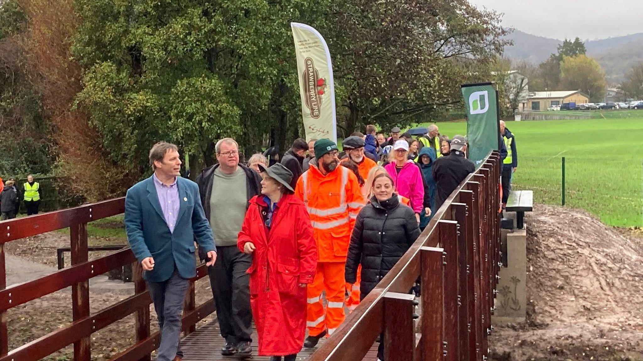 A big group of people, some with high-viz jackets on, walking across a new bridge. The weather is quite wet. There is big banners saying Strawberry Line as they all cross the new bridge.