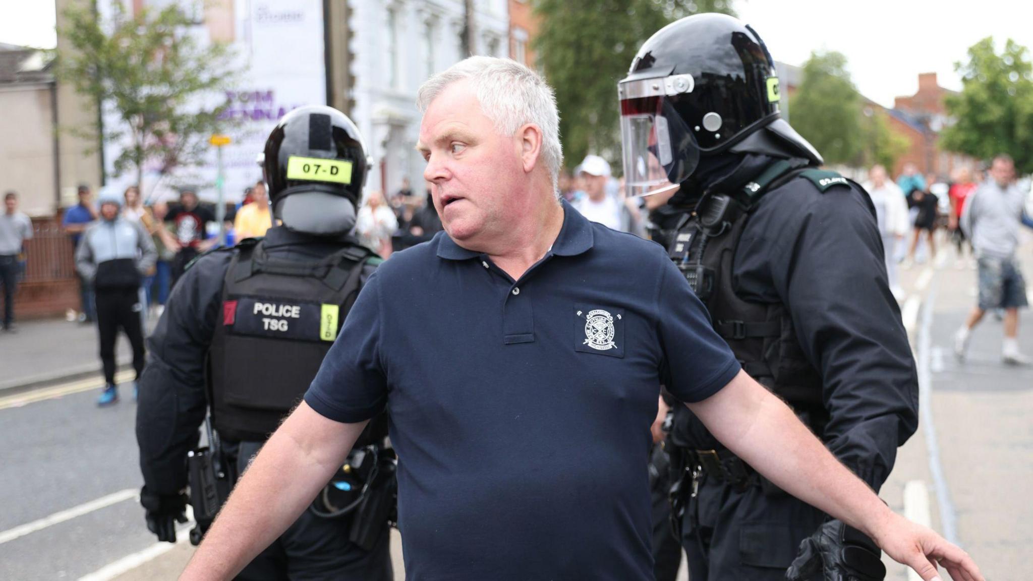 Gerard Rice standing with his arms out with two police officers in riot gear behind him. There is a crowd of protestors out of focus in the background. Mr Rice is wearing a navy polo shirt and he has grey hair.