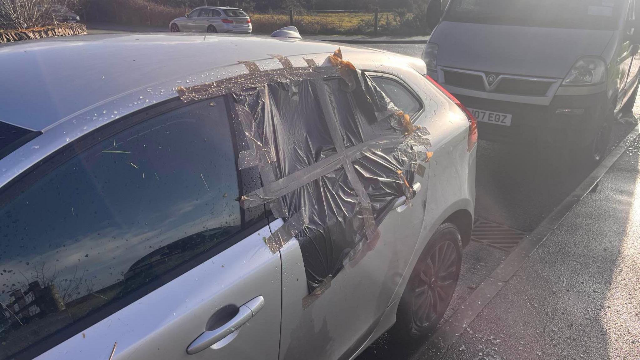 A silver car with a garbage bag on the door after a window was smashed.