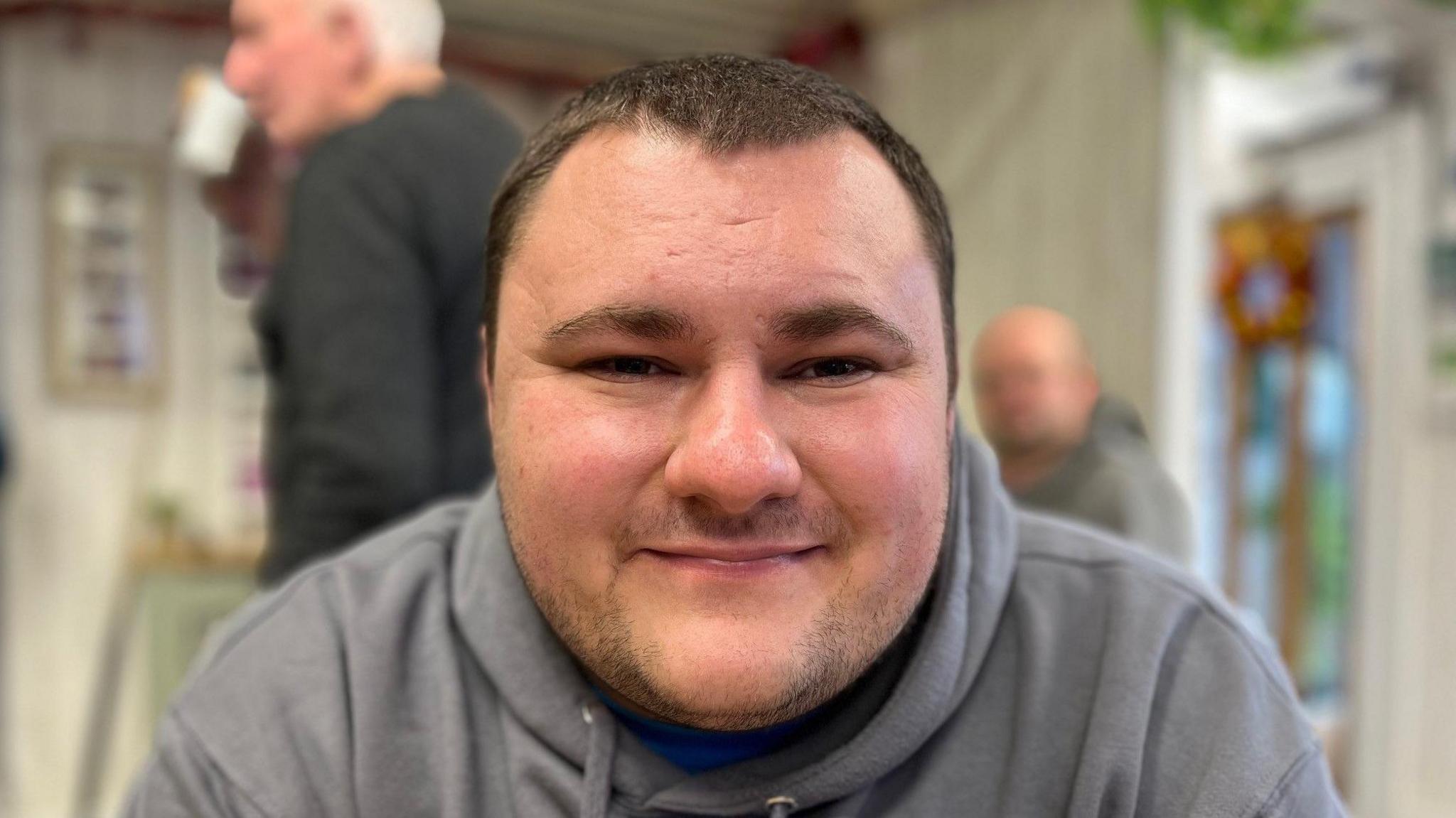 Jay Street is a 26-year-old man with short black hair. This photograph is of his head and shoulders. He's wearing a grey hoodie and is pictured in a café setting in Rhyl, with people mingling behind him.