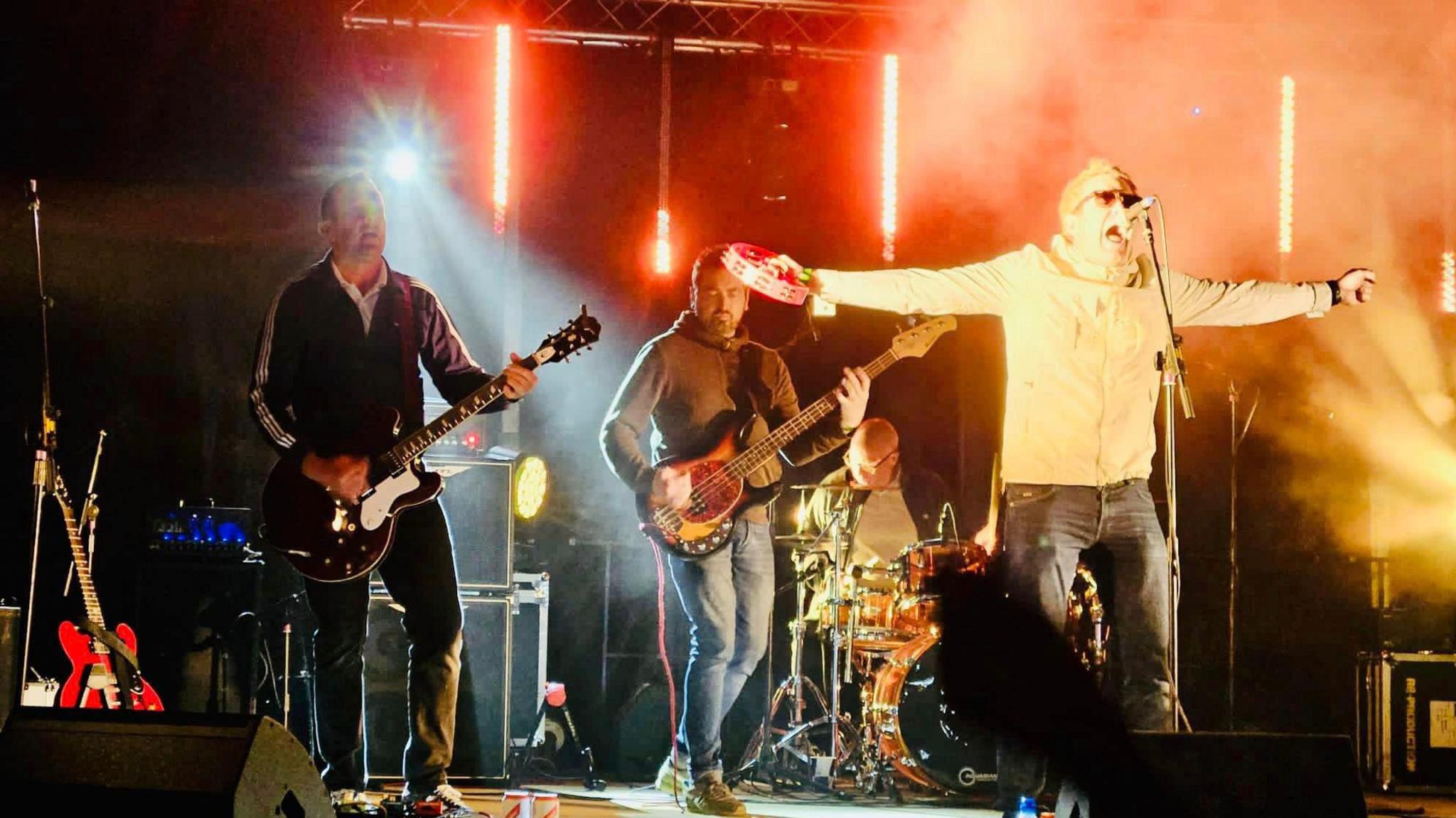 Four men on a lit stage playing a guitar and drums and one at the front of the stage is singing.