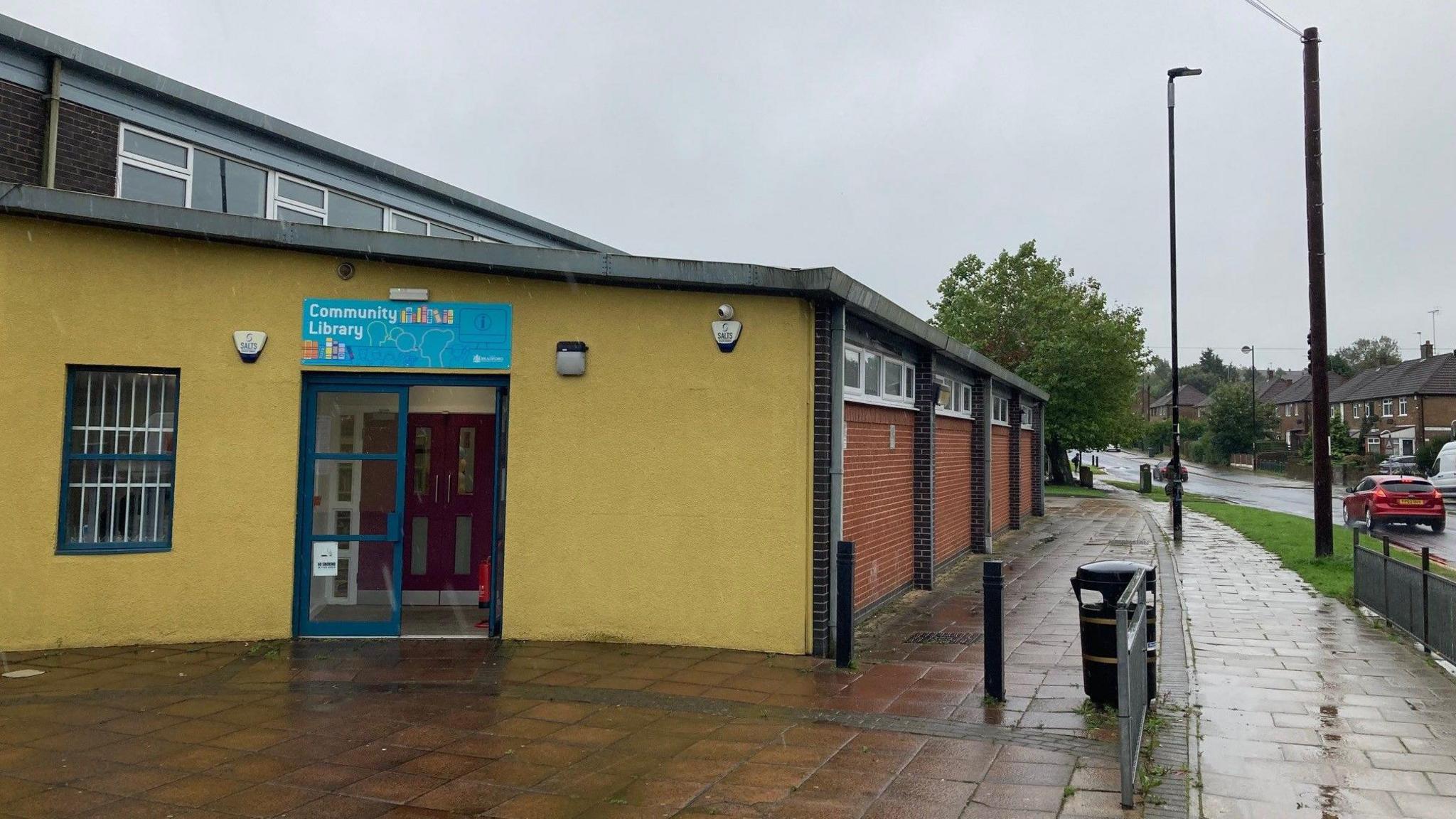 A yellow, pebbledash concrete wall with blue double doors leading into Holmewood Library with a recently rained upon pavement outside.