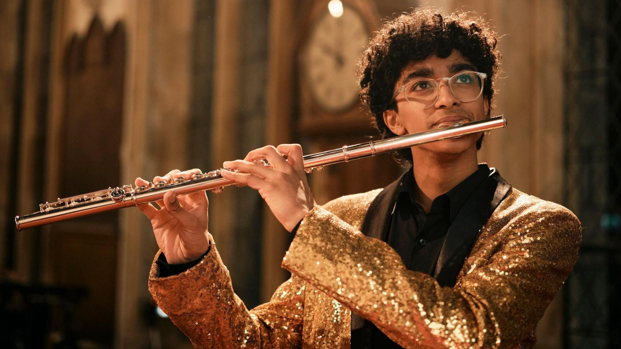 Flute player Cosimo Dhami-Hounsome playing his instrument while wearing a gold sparkly blazer and a black shirt. Cosimo has thick curly black hair and is wearing clear-rimmed glasses. Cosimo is looking up towards the roof of the building.