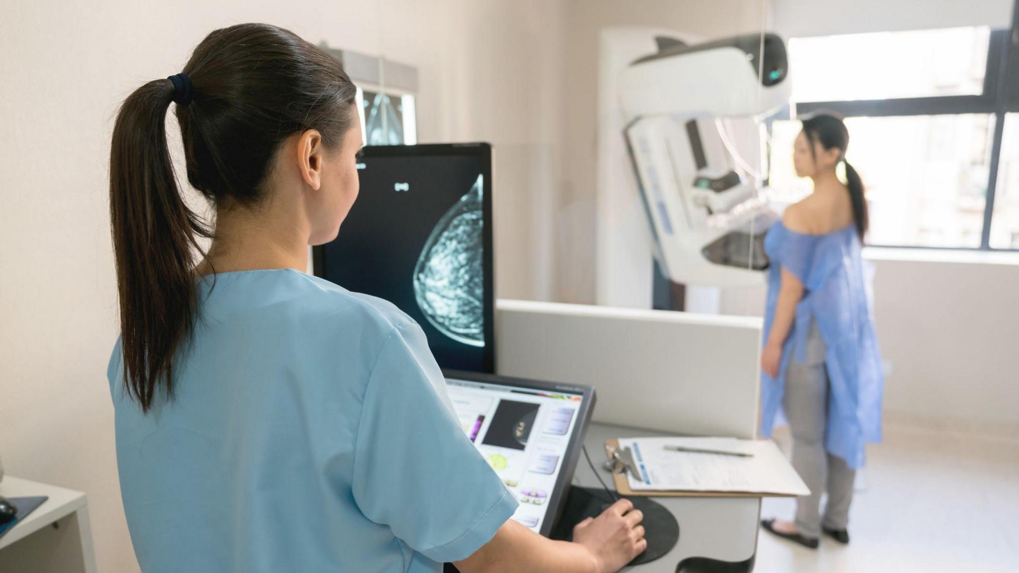 A doctor wearing blue scrubs is checking a scan while a woman is standing in the background wearing blue clothing protection receiving a scan.