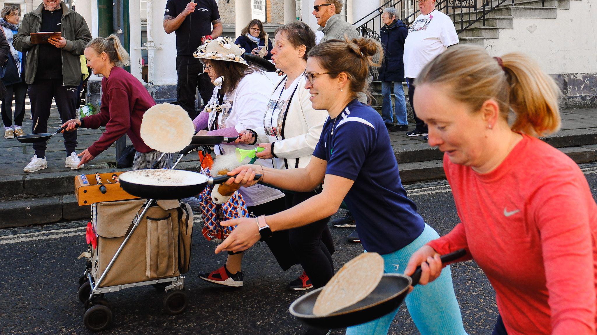 Five women are lined up in the road taking part in a pancake race. They are all concentrating on trying to flip their pancake in their frying pan, while running through the street as onlookers watch.