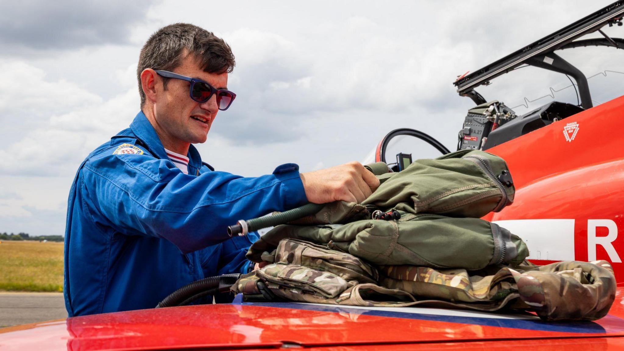 A man dressed in blue overalls is placing air crew kit on the wing of a red plane. It is a sunny day and he is wearing dark glasses and has short, dark hair.
