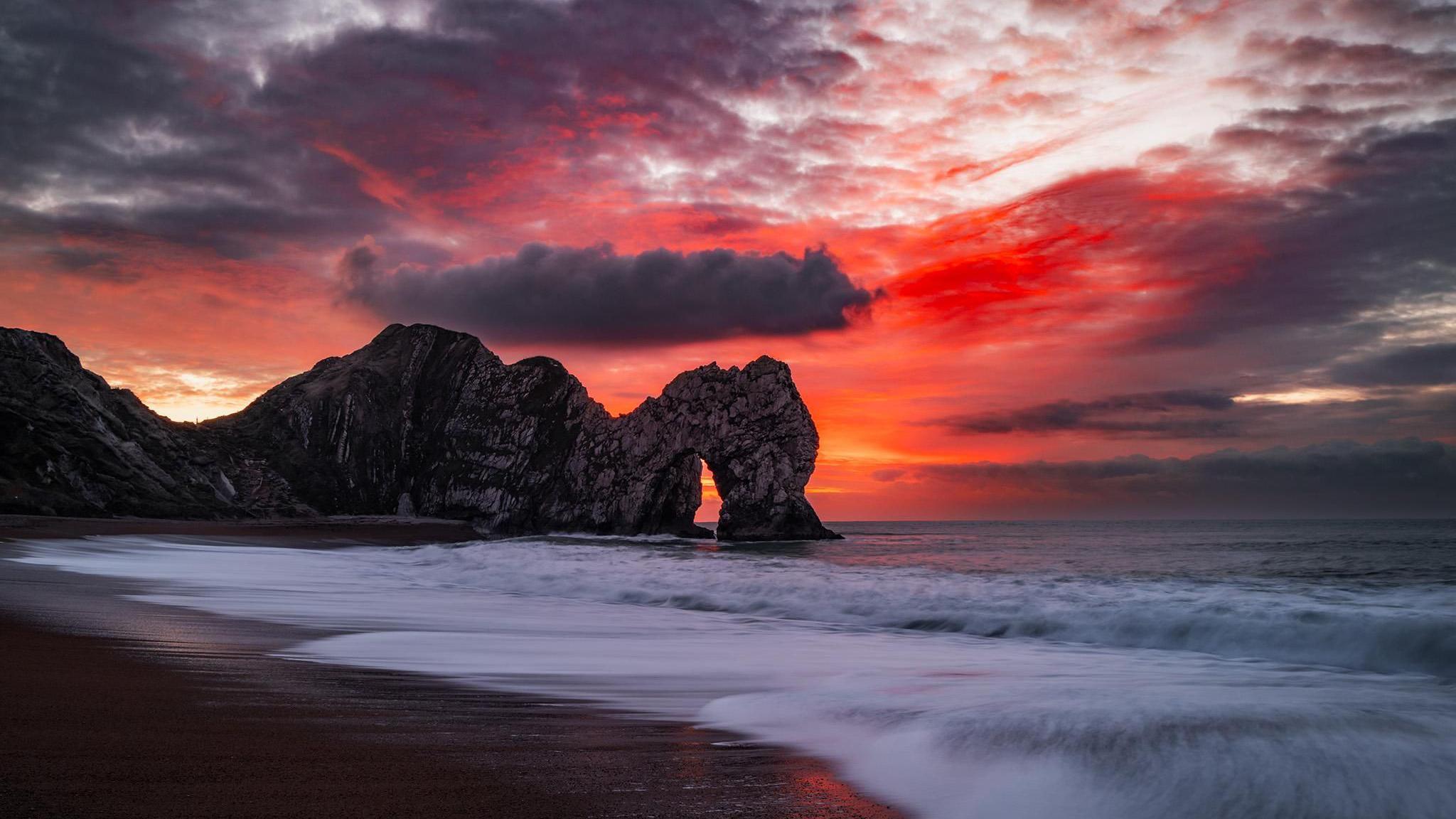 The rock arch of the Durdle Door cliffs is illuminated by a bright red sky. There are waves washing up on the beach and dark clouds in the sky. 