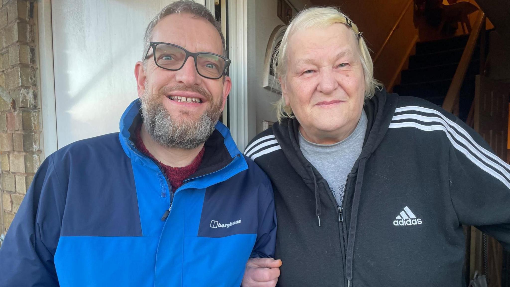 A man in a blue winter jacket and an older lady in a sport coat smile arm in arm