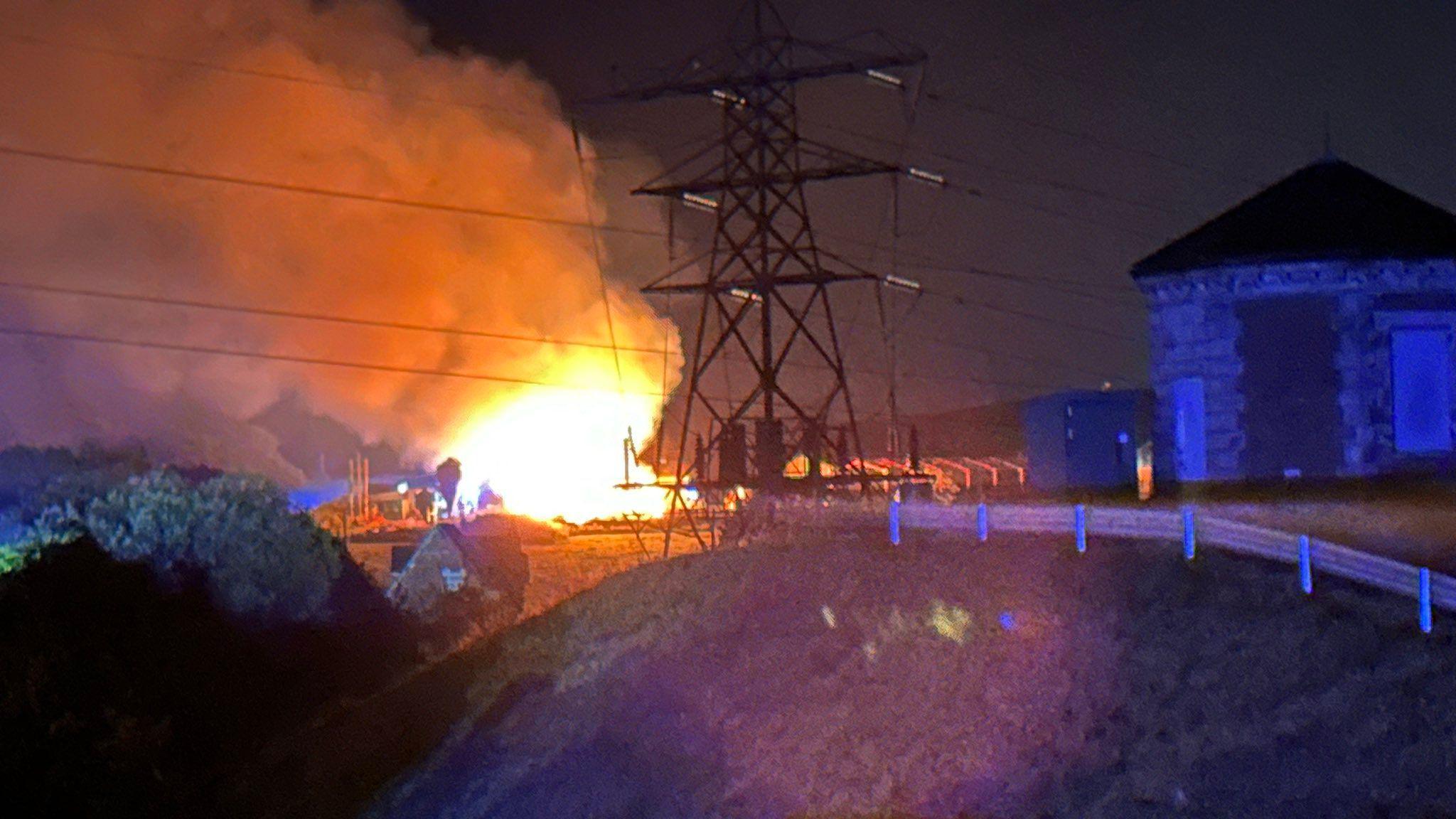 A large fire can be seen in the background with a telegraph pylon in the middle distance and a fence as well as a building illuminated by blue light