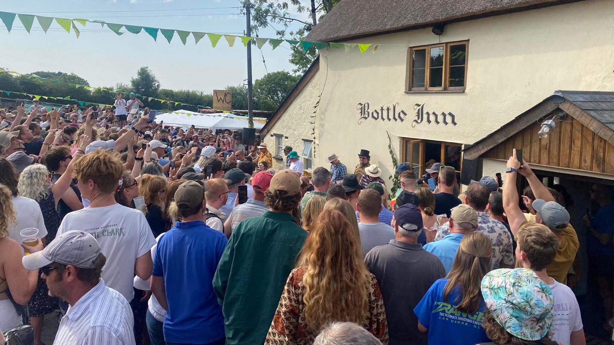 Crowds of people, many holding phones aloft, outside the cream-coloured, thatched Bottle Inn