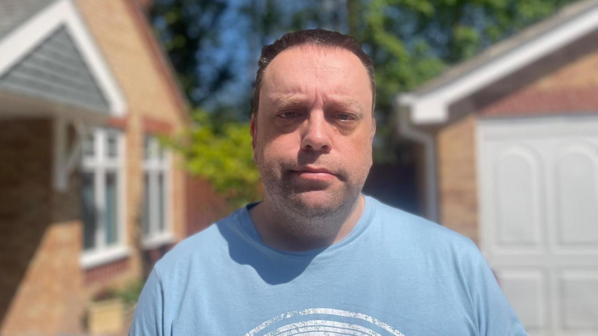 A middle aged man in a blue t-shirt looks dejected towards the camera outside a red brick house