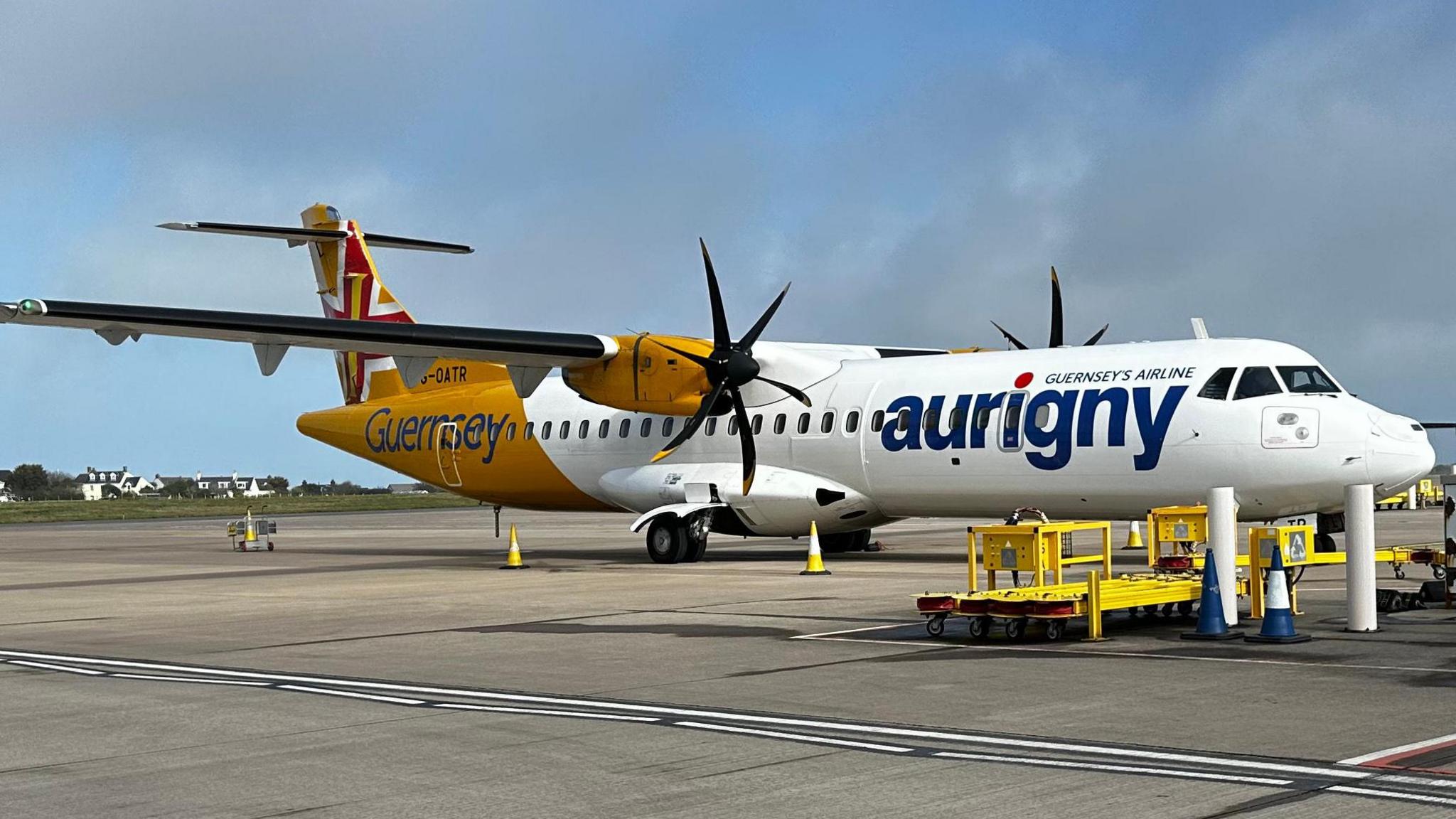An Aurigny aircraft with its yellow and white livery. It has two propellers. The aircraft is on a runway at an airport.