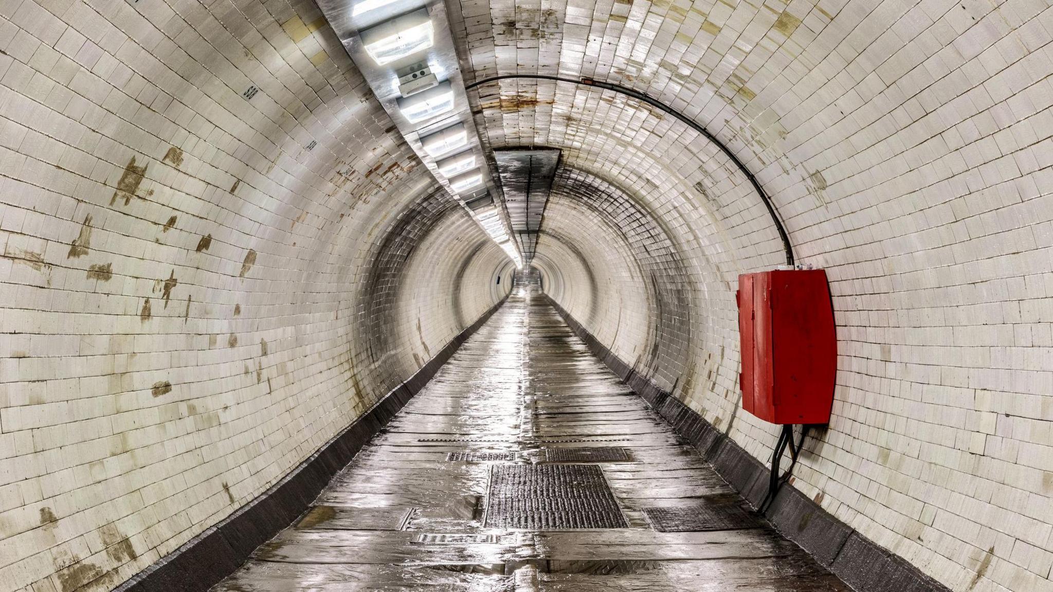 A white tiled tunnel with a red box attached to the wall on the right hand side. The tunnel is empty and is lit by overhead fluorescent lights. 