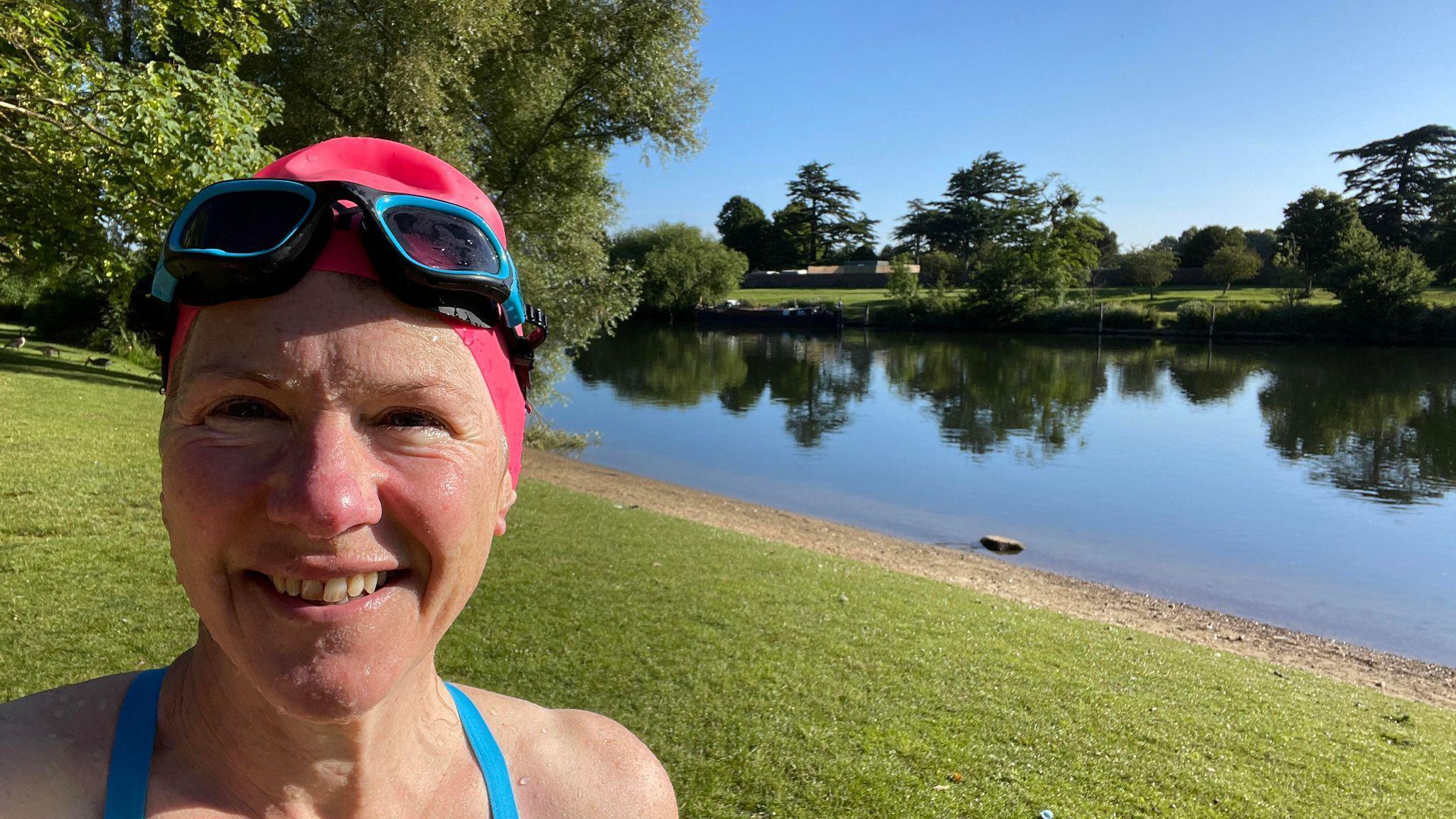 A woman with a swimming hat and goggles smiles widely up close to the camera with a sunny river bank in the background.