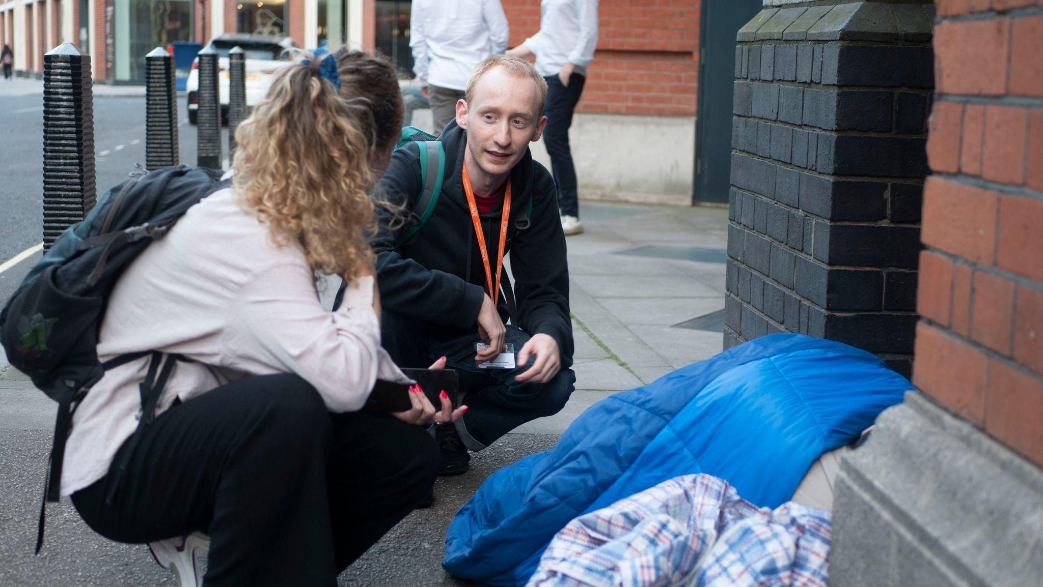A man and a woman crouch down on the street next to a person in a blue sleeping bag.
