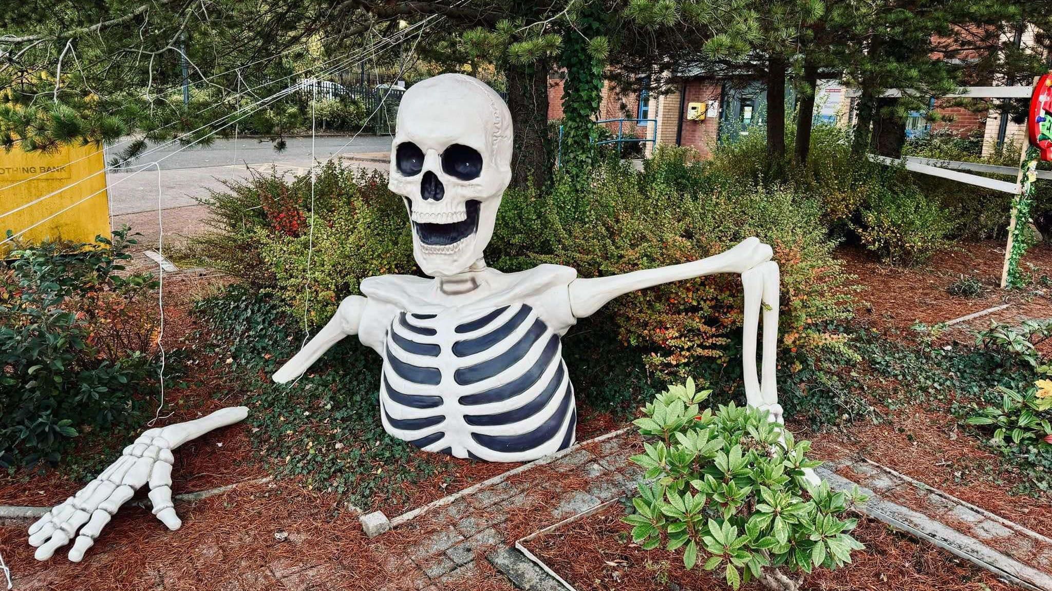 A large white skeleton torso in the ground with bushes, trees and spider webs in the background.