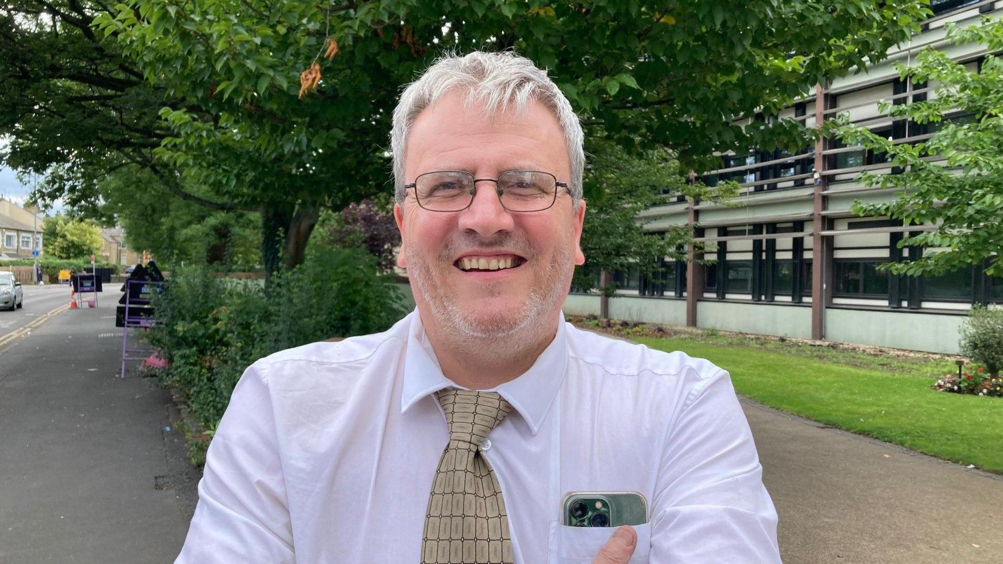 A smiling man with white hair wearing glasses and a white shirt with a yellow, patterned tie.
