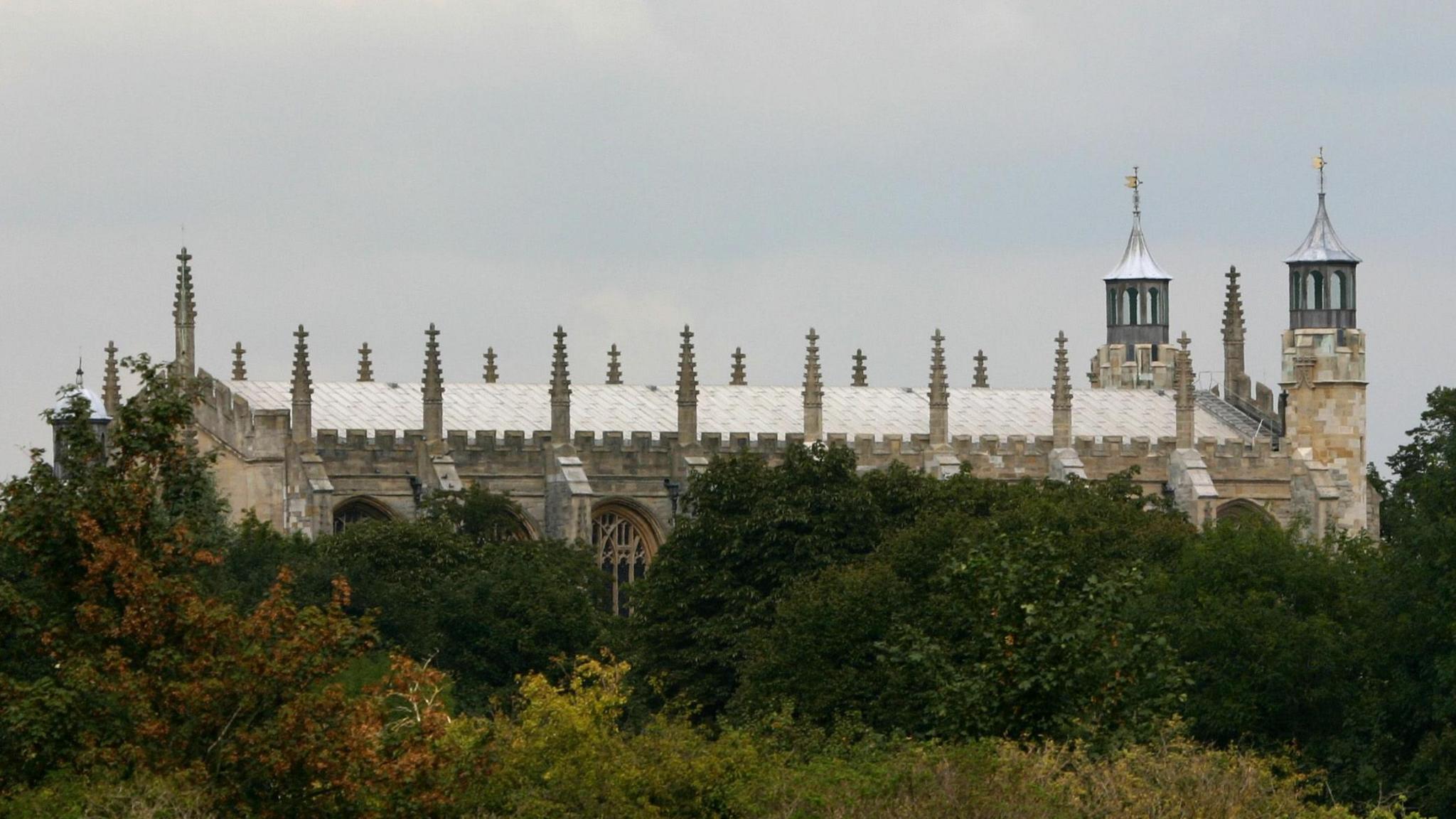 The chapel of Eton College, seen from the Thames at Windsor, Berkshire. It has a giant roof with two spires at one side. It pokes out of greenery.