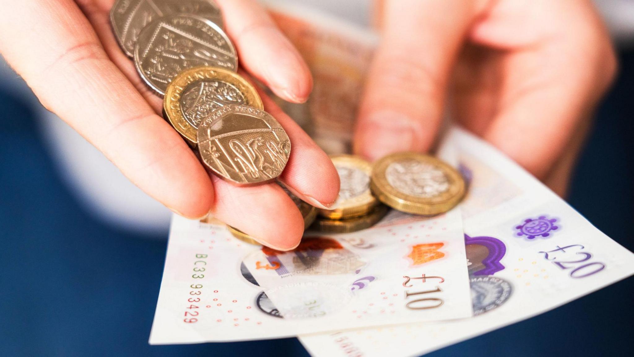 A pair of hands holding UK coins and a couple of crisp banknotes.