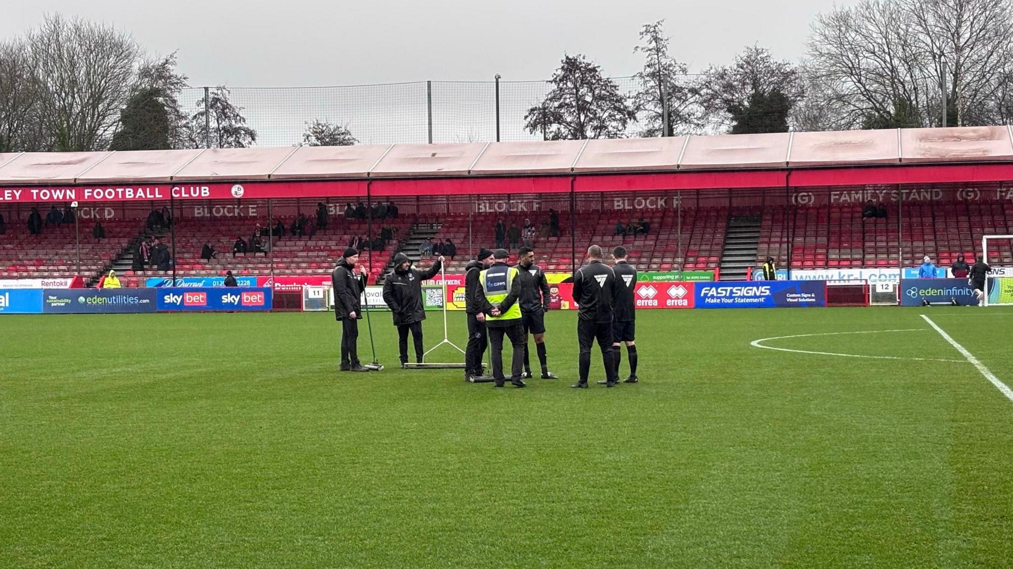 Six people in all black clothing stood in the middle of a football pitch. In the background are red seating stands