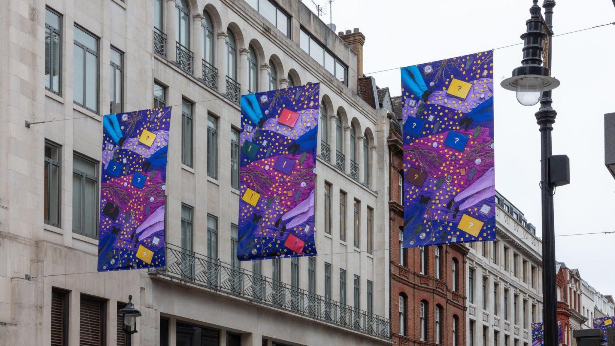 Three banners hanging across a street, they are purple, pink, yellow and blue. 