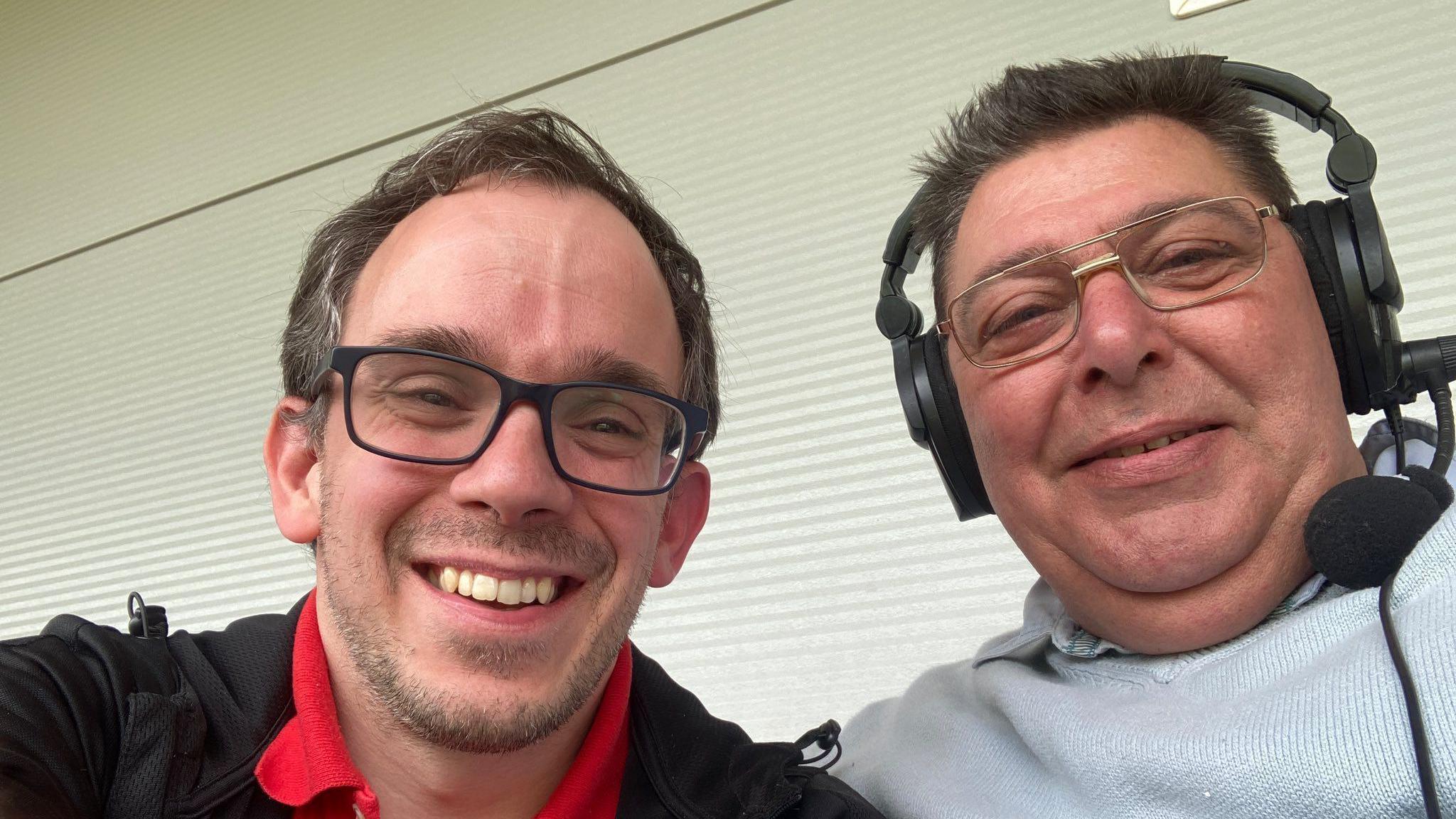 Two smiling men in glasses, one is wearing a red polo shirt and a black jacket while the other is wearing a grey jumper and had a headset on